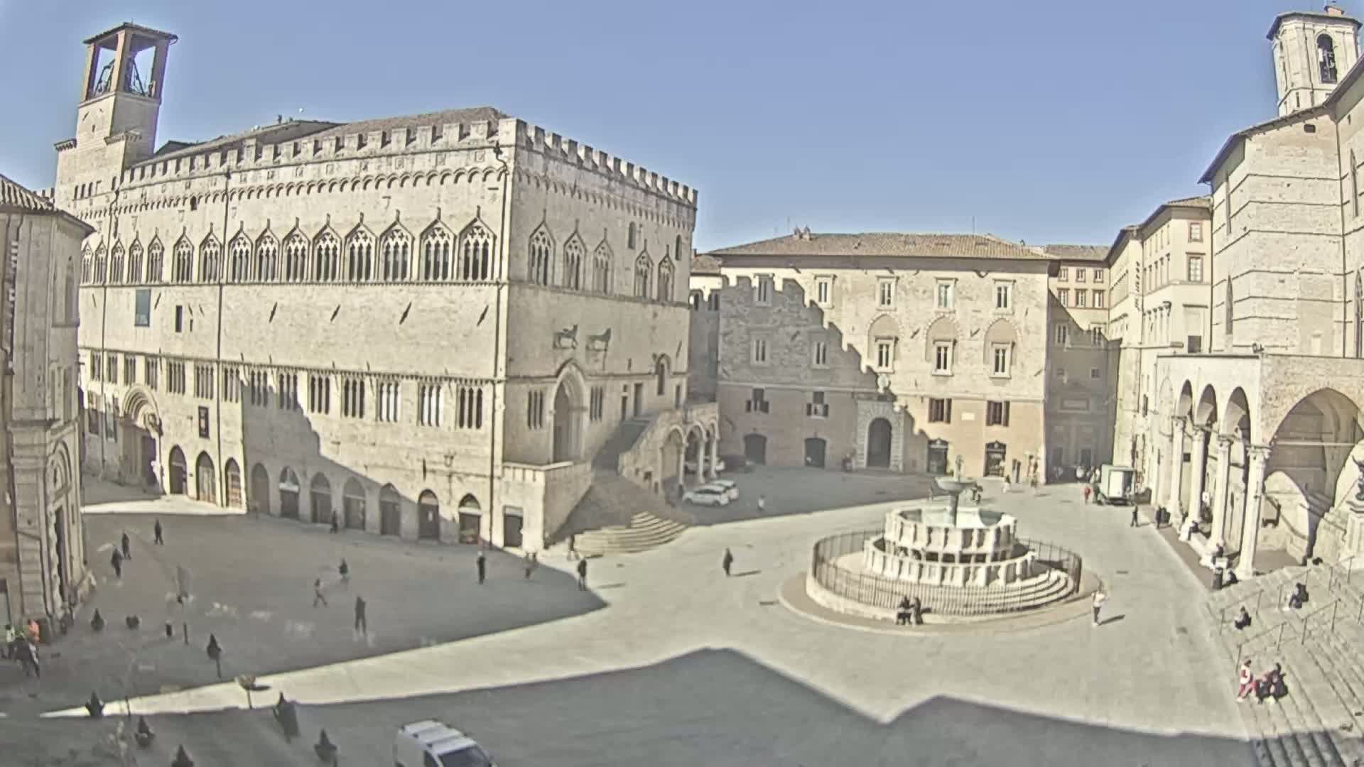 A large, sunlit town square features a fountain, several stone buildings, and a scattering of people.