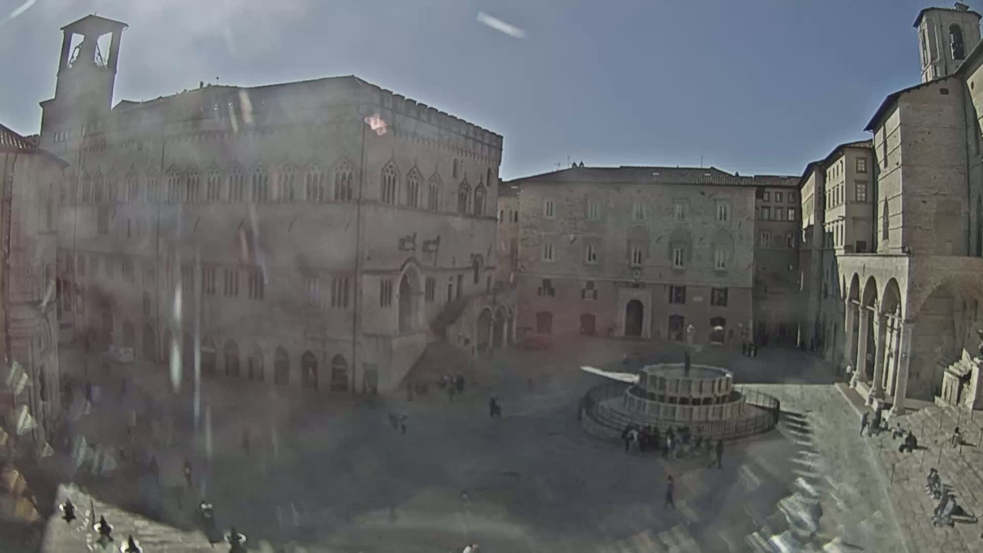 A sunny day reveals a town square surrounded by historic stone buildings, a central fountain, and a few scattered people.