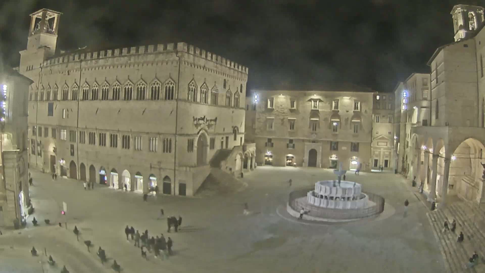A nighttime view of a large, empty town square with several historic stone buildings surrounding a central fountain, under dark, clear skies.