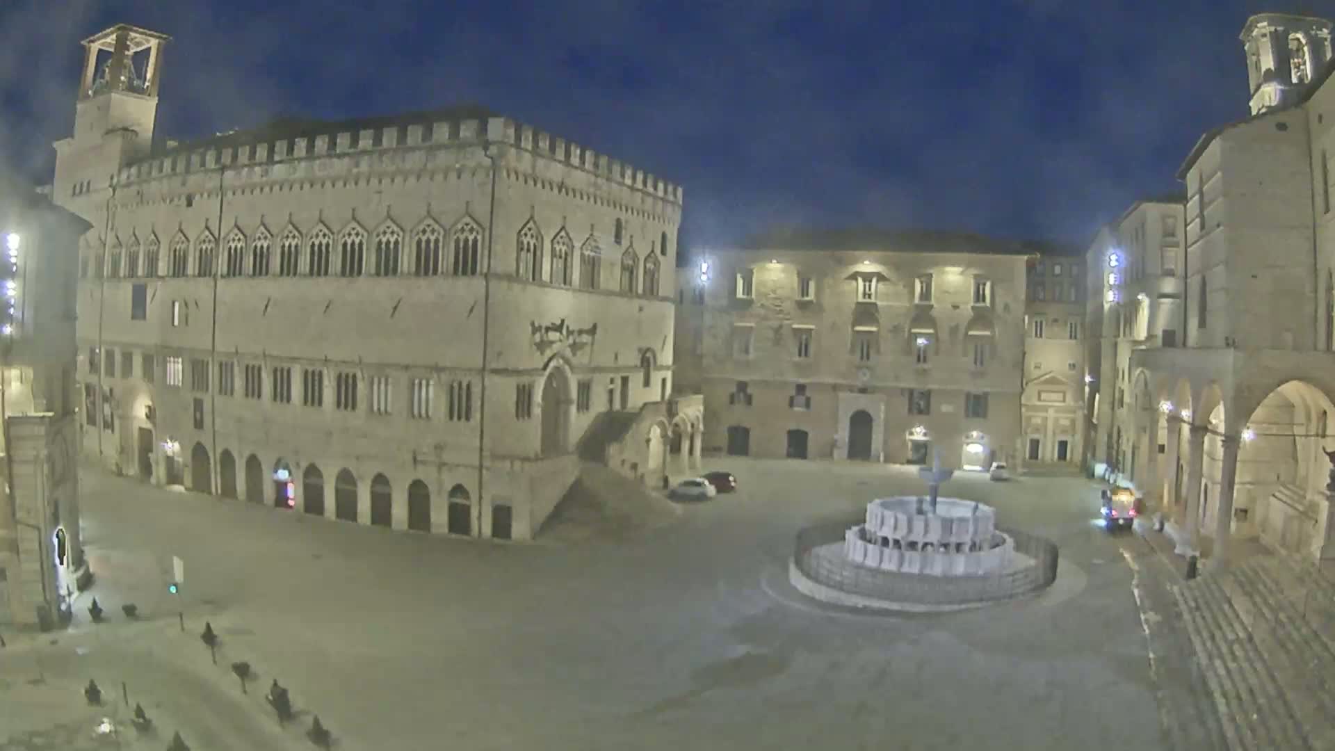 A mostly empty town square at night, featuring a large stone building with arched entryways and a fountain in the center, under a dark, clear sky.