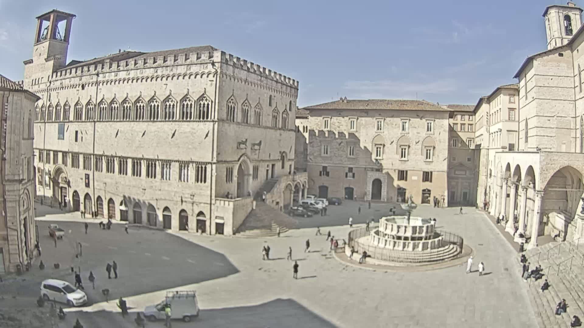 A large, paved town square, under sunny skies, is surrounded by several aged stone buildings, one prominently featuring a staircase, and a central fountain.