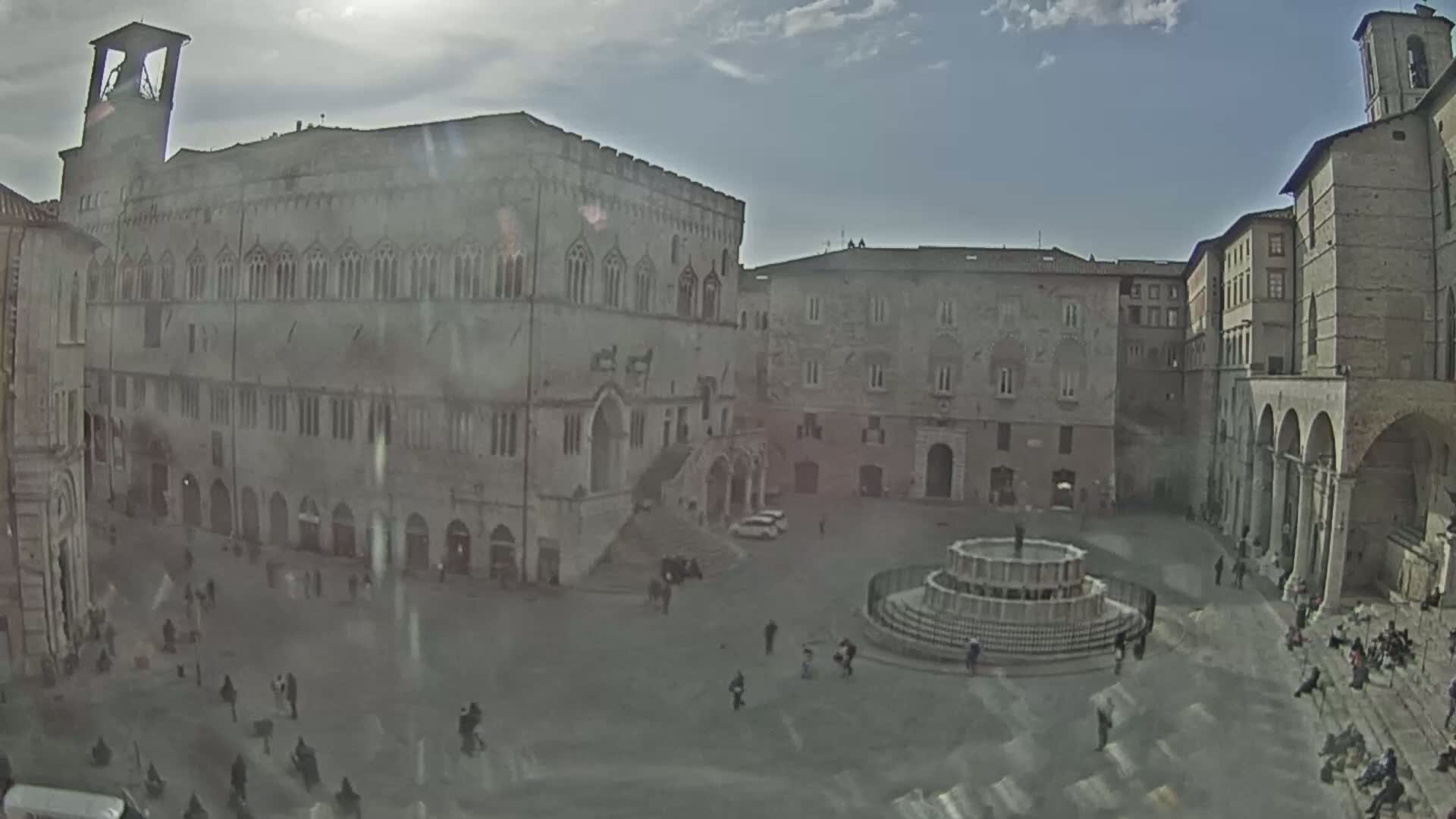 A large, stone public square with a fountain in the center is surrounded by historic buildings under a partly cloudy sky.