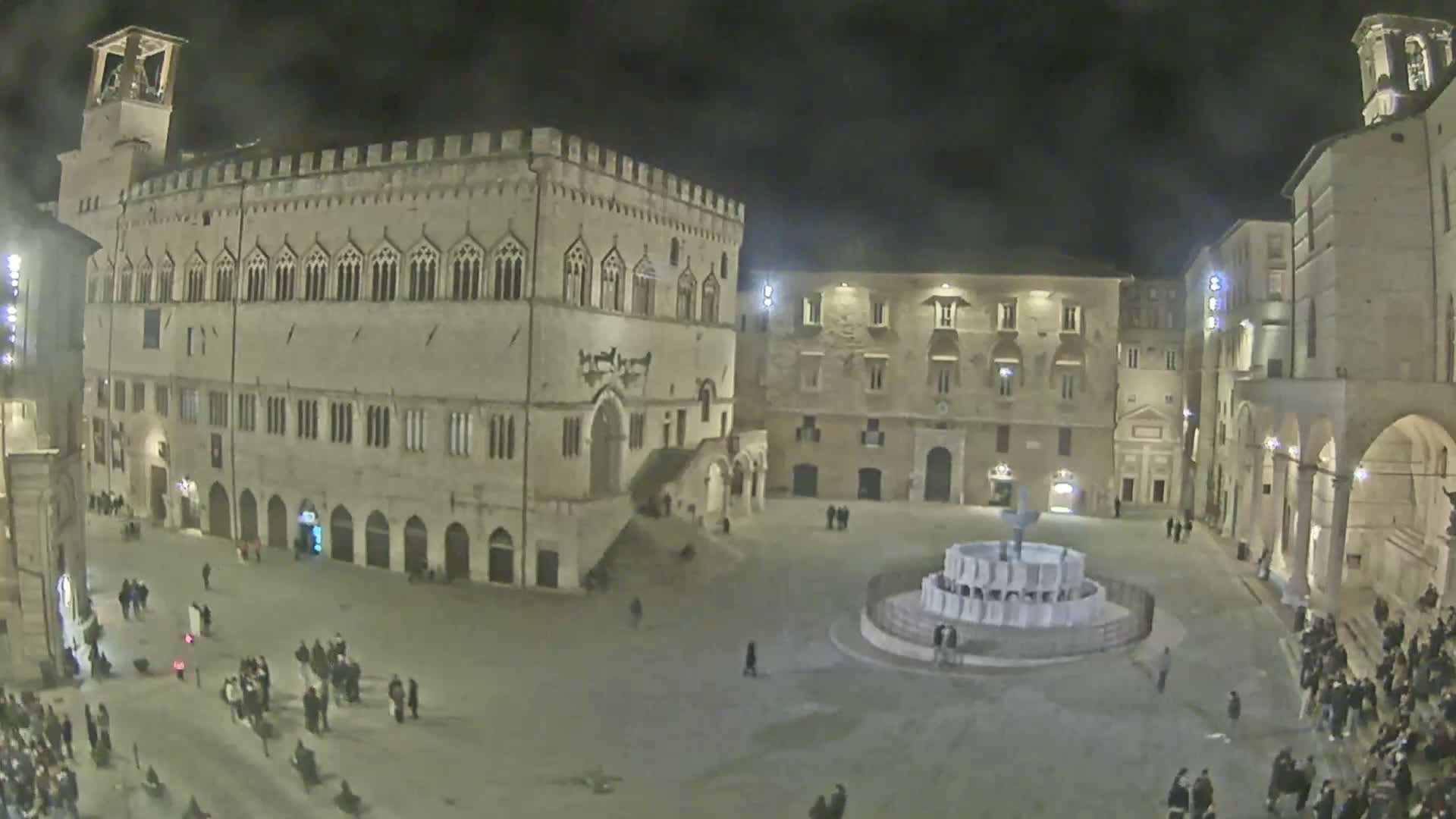 A nighttime, high-angle view shows a town square with several large buildings surrounding a fountain, and many people are scattered throughout the square.
