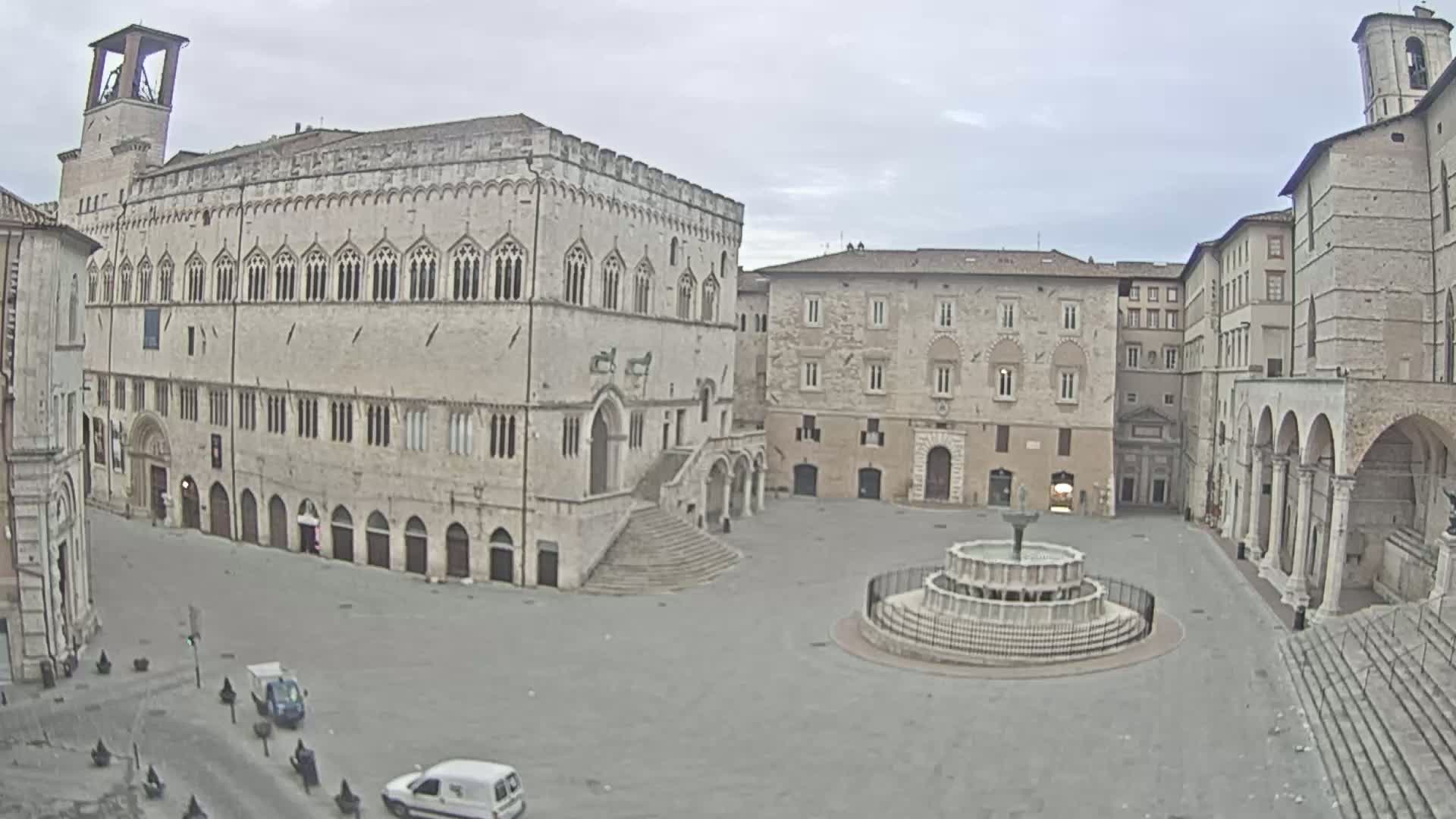 A large, empty plaza with a fountain in the center is surrounded by several aged stone buildings under an overcast sky.