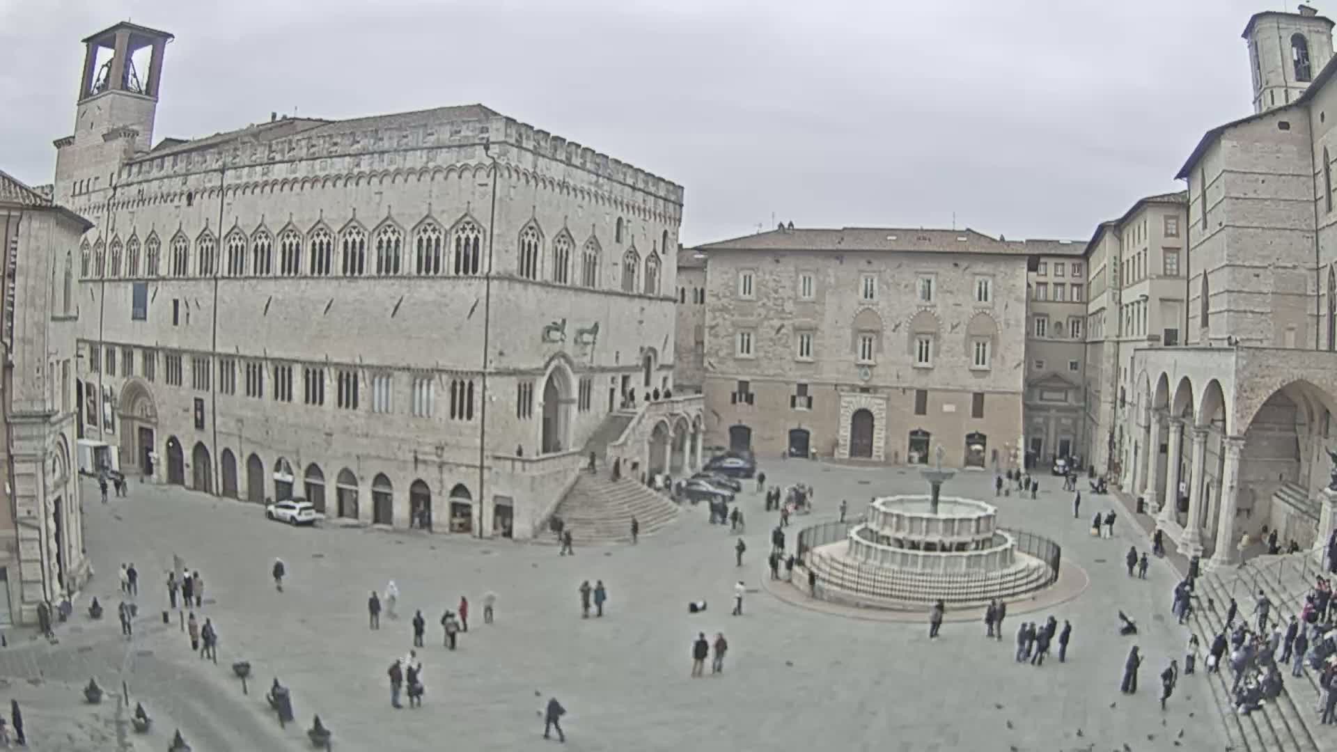 A large town square with stone buildings and a fountain, under an overcast sky.