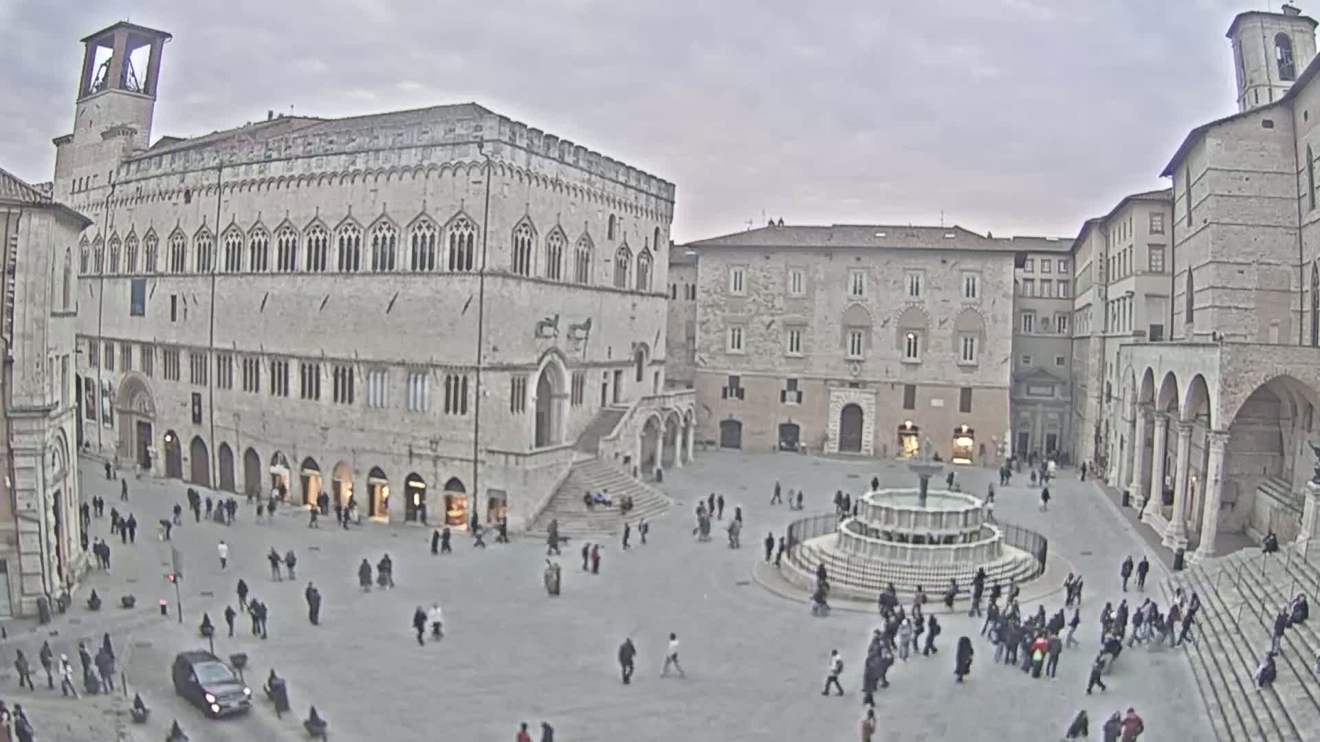 A large town square, under an overcast sky, features a central fountain and is surrounded by several historic stone buildings.