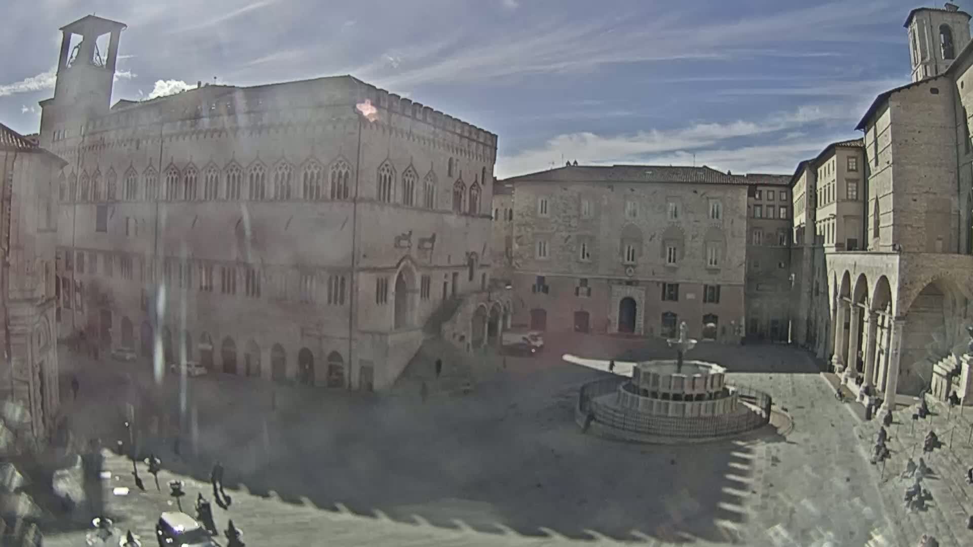 A town square with a fountain is surrounded by several aged stone buildings under a partly sunny sky.