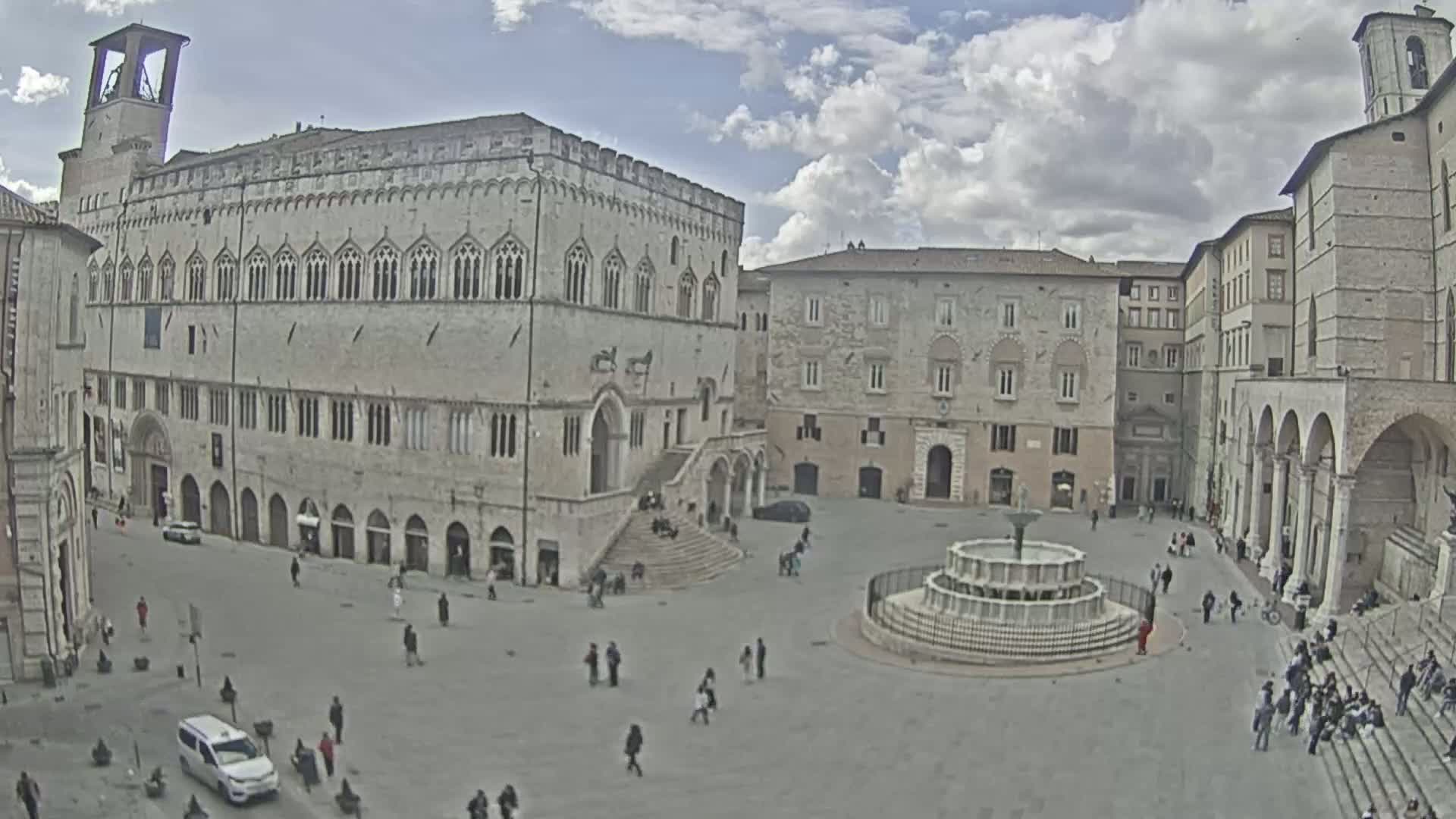 A large town square with several stone buildings, a fountain, and scattered pedestrians under a partly cloudy sky.
