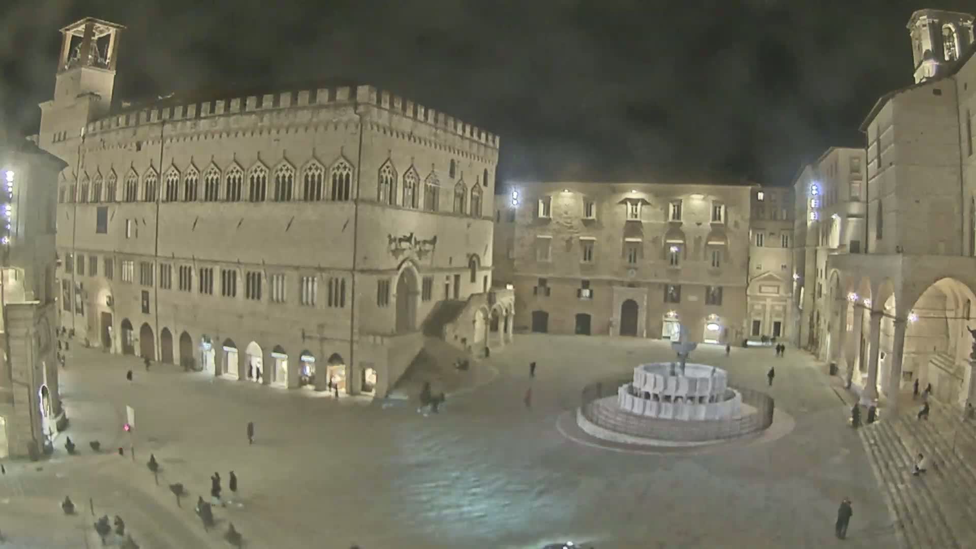 A large, stone plaza at night, featuring a central fountain and several historic buildings, is sparsely populated with a few pedestrians.