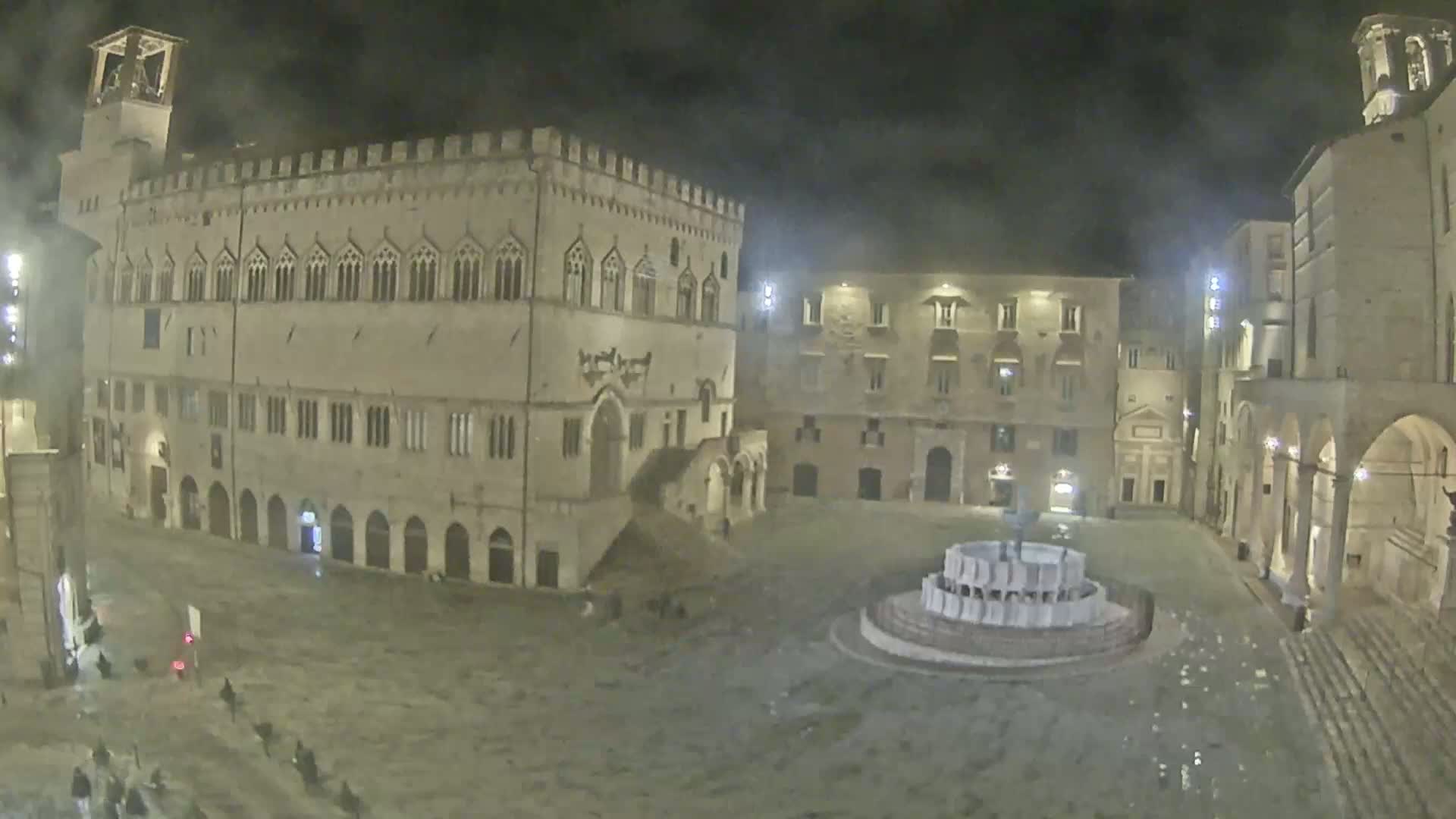 A nighttime, wet, stone piazza is centered on a fountain, surrounded by several large, historic buildings.