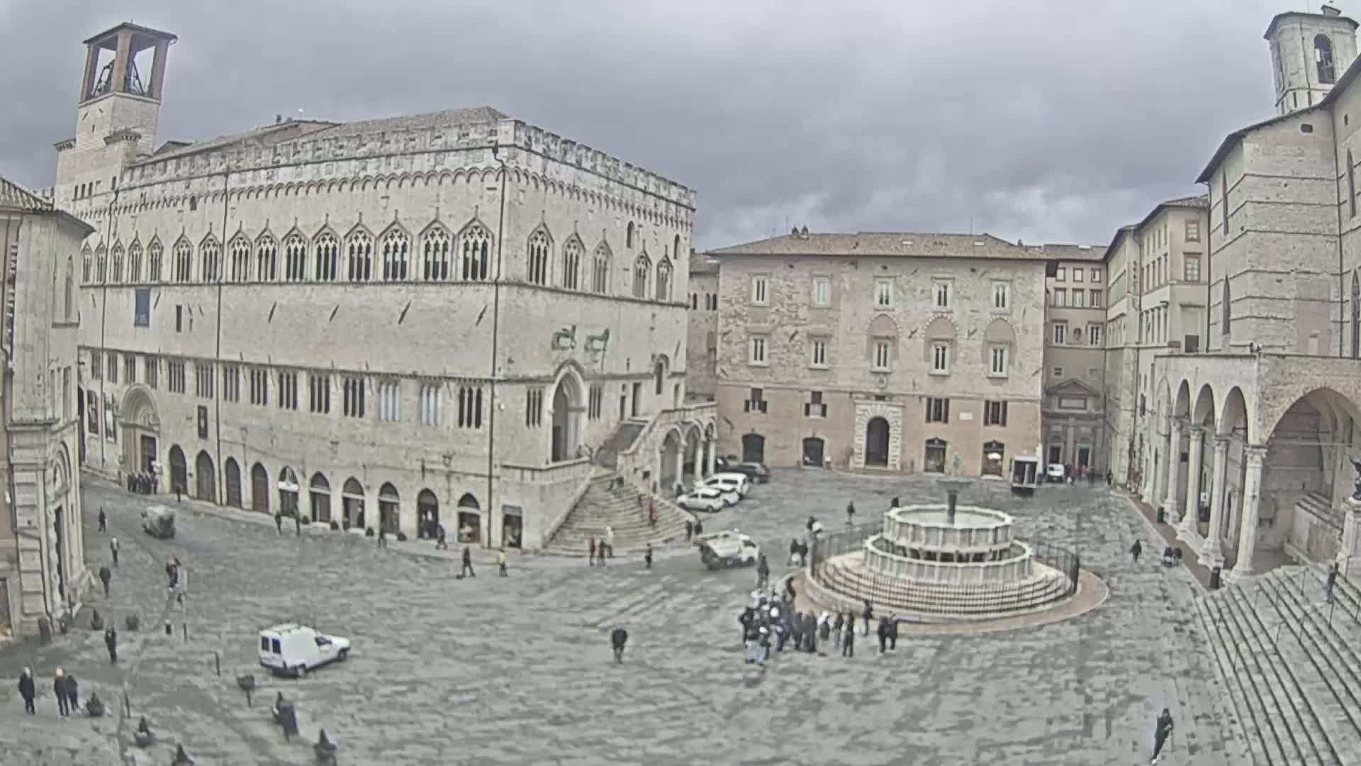 A large, stone plaza with a fountain at its center is surrounded by historic buildings under an overcast sky.