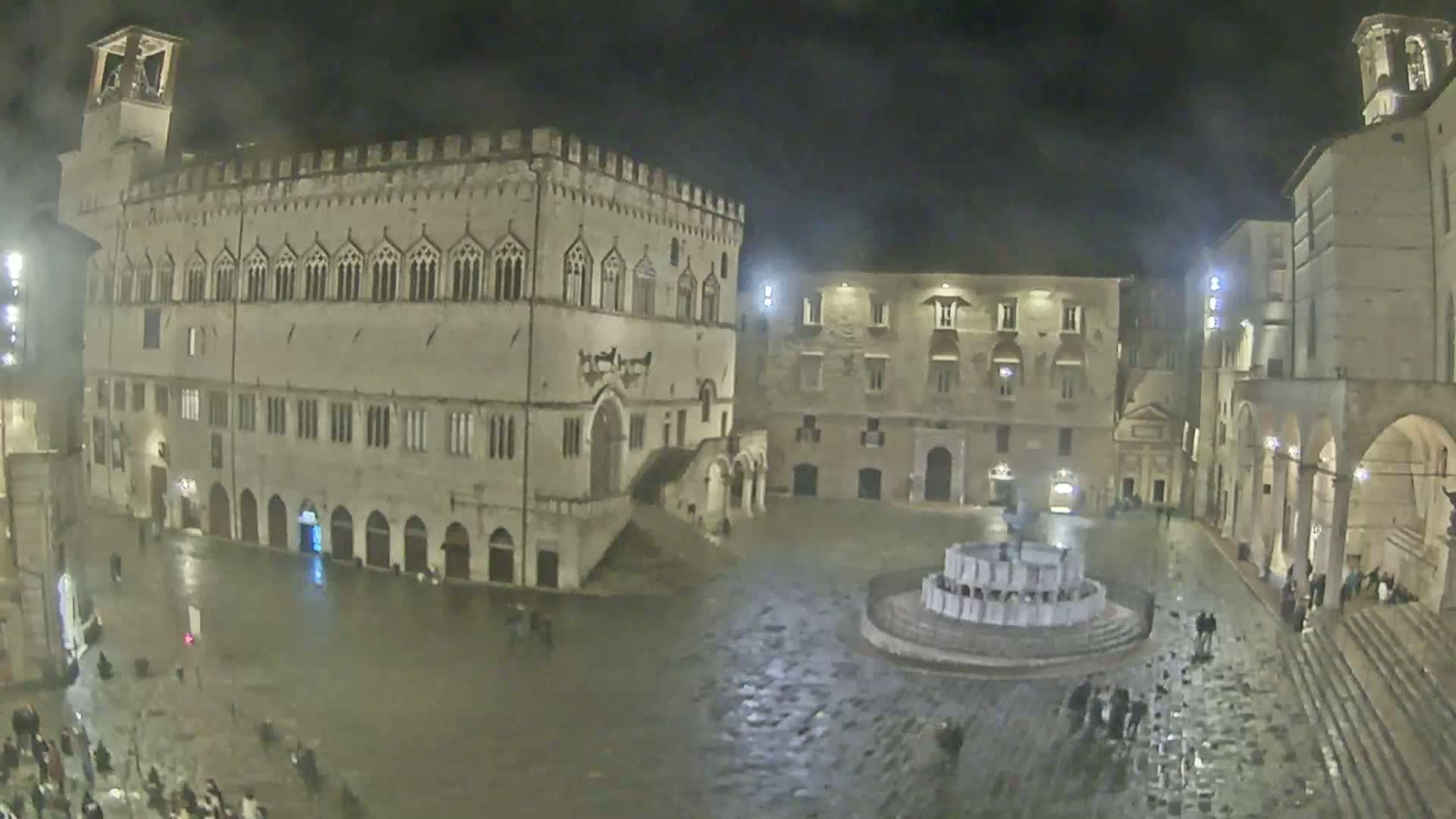A wet, nighttime view of a town square surrounded by historic stone buildings, centered on a fountain.