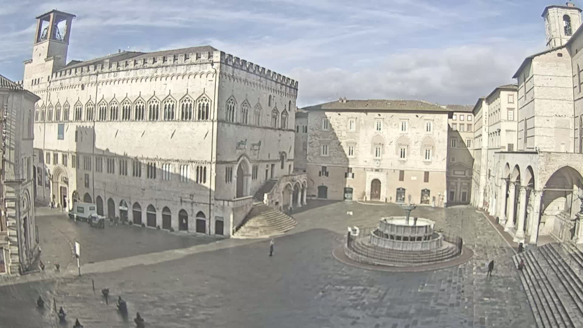 A large, mostly empty town square with a central fountain is surrounded by several historic stone buildings under a partly cloudy sky.