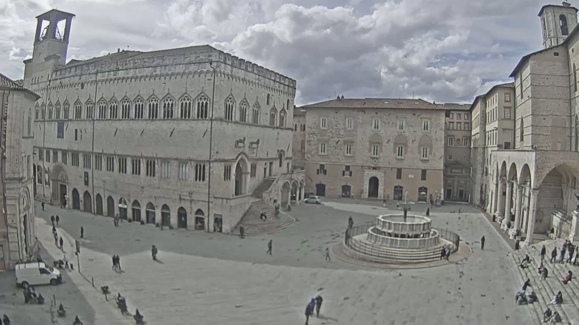 A large town square, under a cloudy sky, features a central fountain and is surrounded by several large, historic stone buildings.