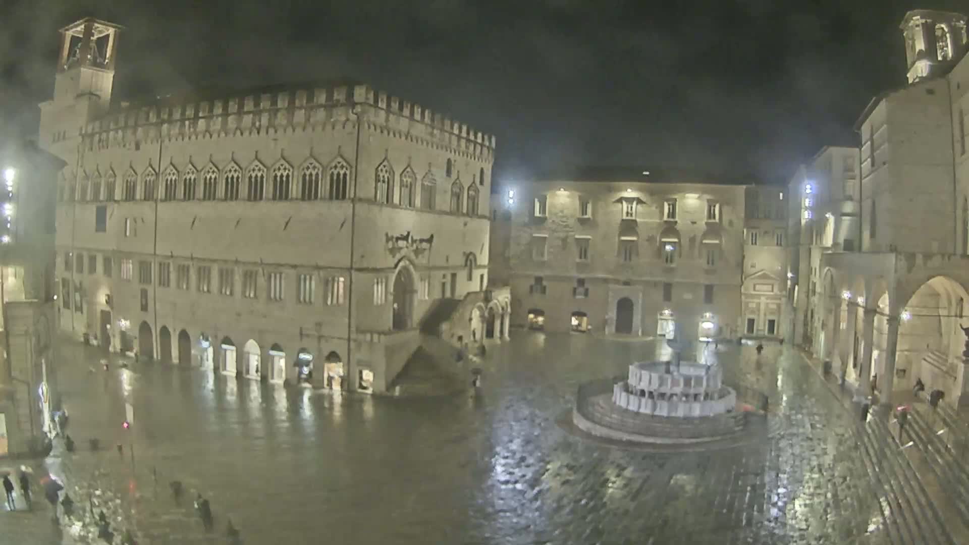 A large, rain-soaked town square at night, surrounded by historic stone buildings, features a central fountain.