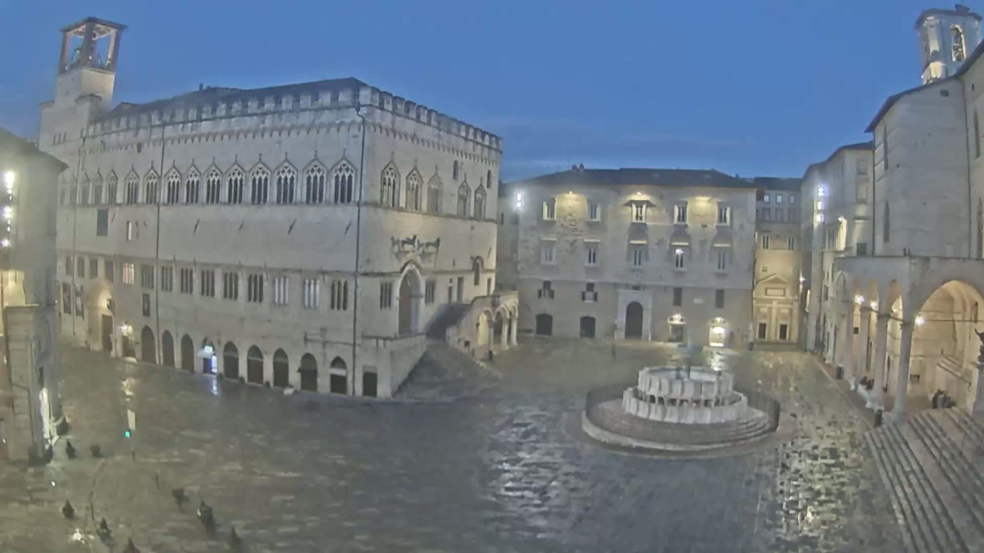 A large, empty town square at night is wet from rain, featuring a fountain in the center and several large stone buildings surrounding it.