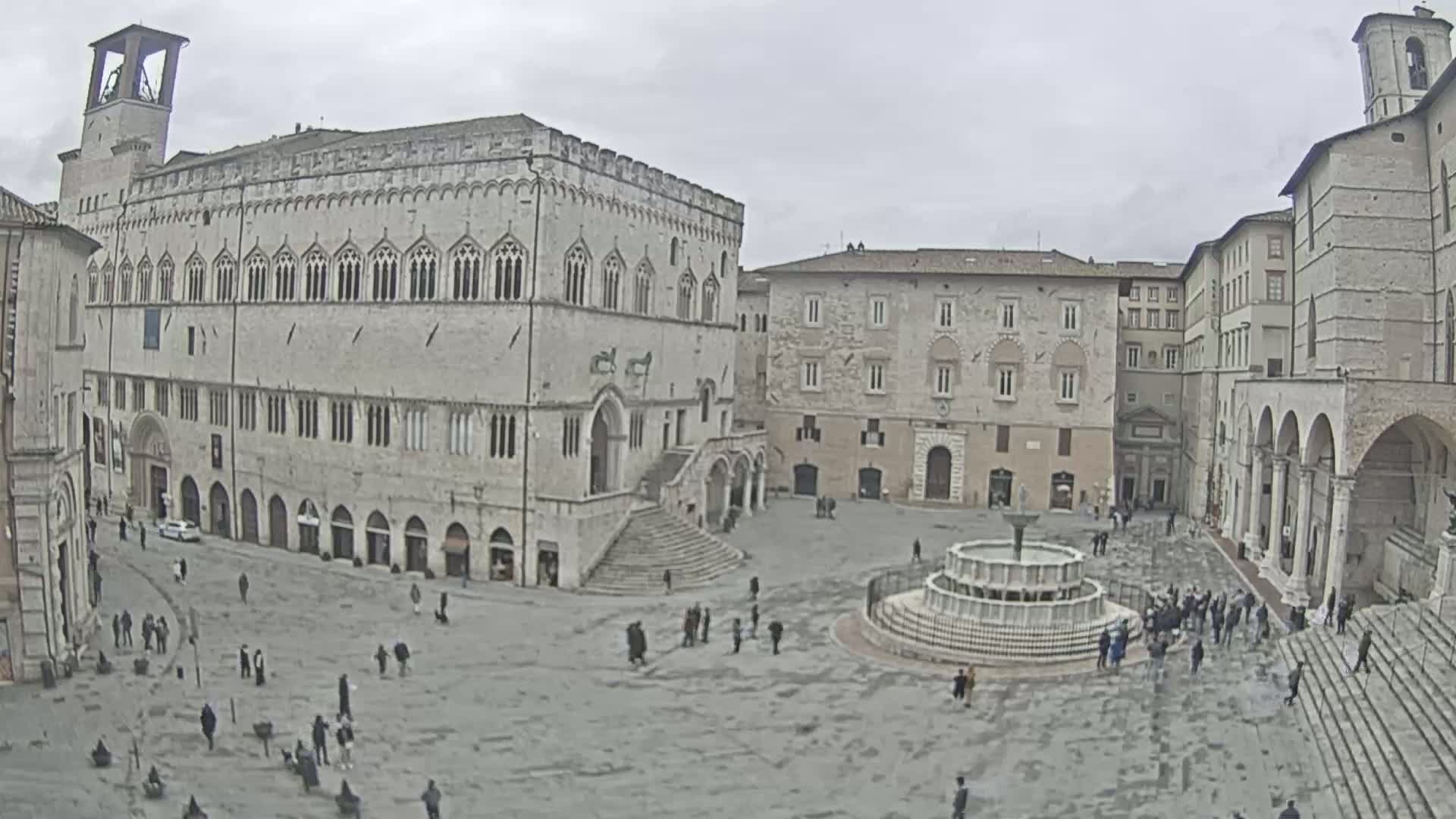 A large, mostly empty town square is surrounded by ancient stone buildings under an overcast sky, featuring a central fountain.
