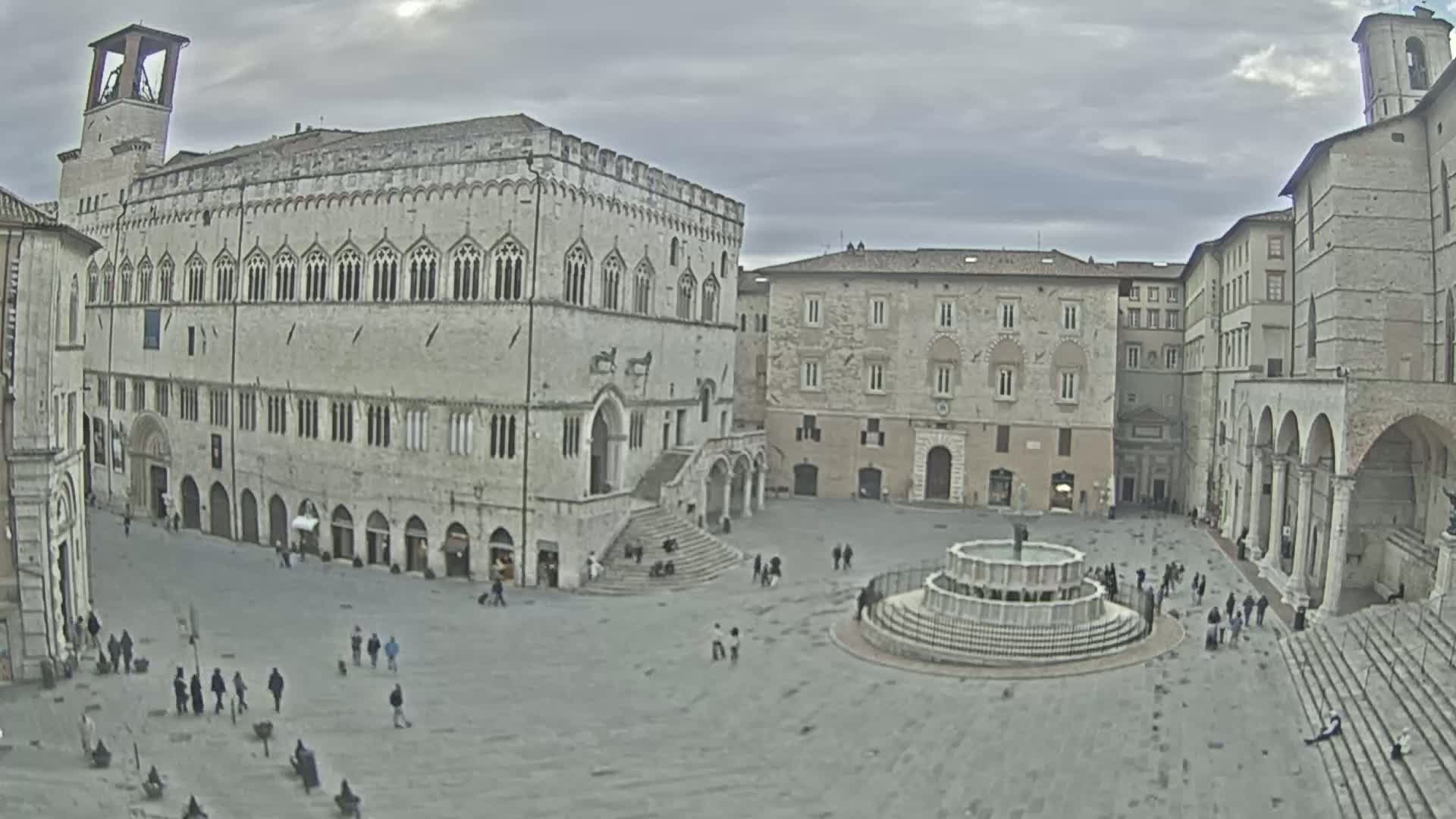 A large, stone plaza with a fountain is surrounded by several multi-story buildings under a cloudy sky.