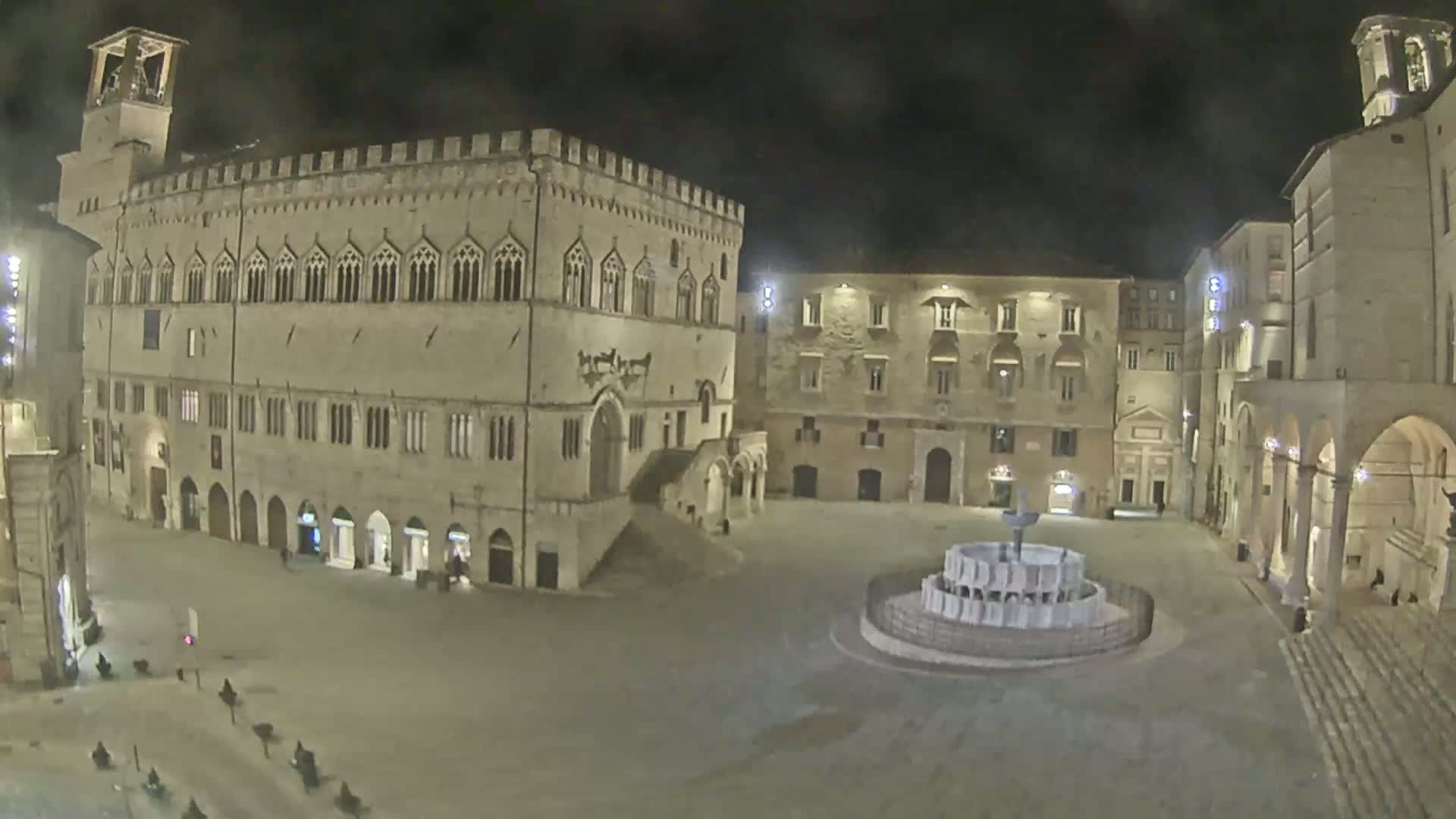 A large, empty plaza at night is surrounded by historic stone buildings, with a fountain in the center.