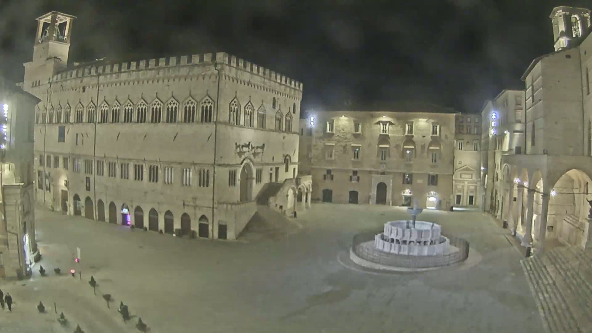 A nighttime view of a large, mostly empty plaza with a fountain in the center, surrounded by historic stone buildings.