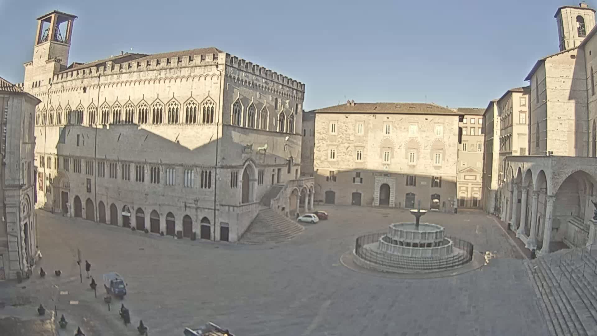 A large, mostly empty town square is surrounded by historic stone buildings under a clear, sunny sky; a fountain sits in the center of the square.