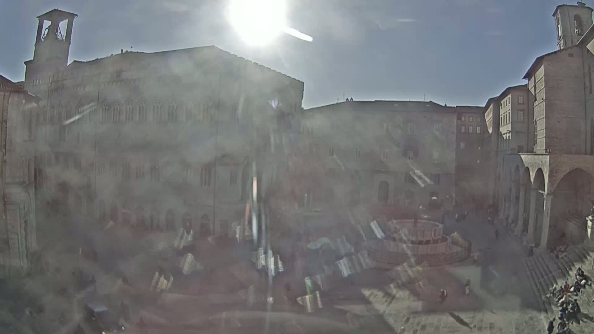 A sun-drenched town square, featuring several historic stone buildings and a central fountain, is populated with a few people on a bright, sunny day.