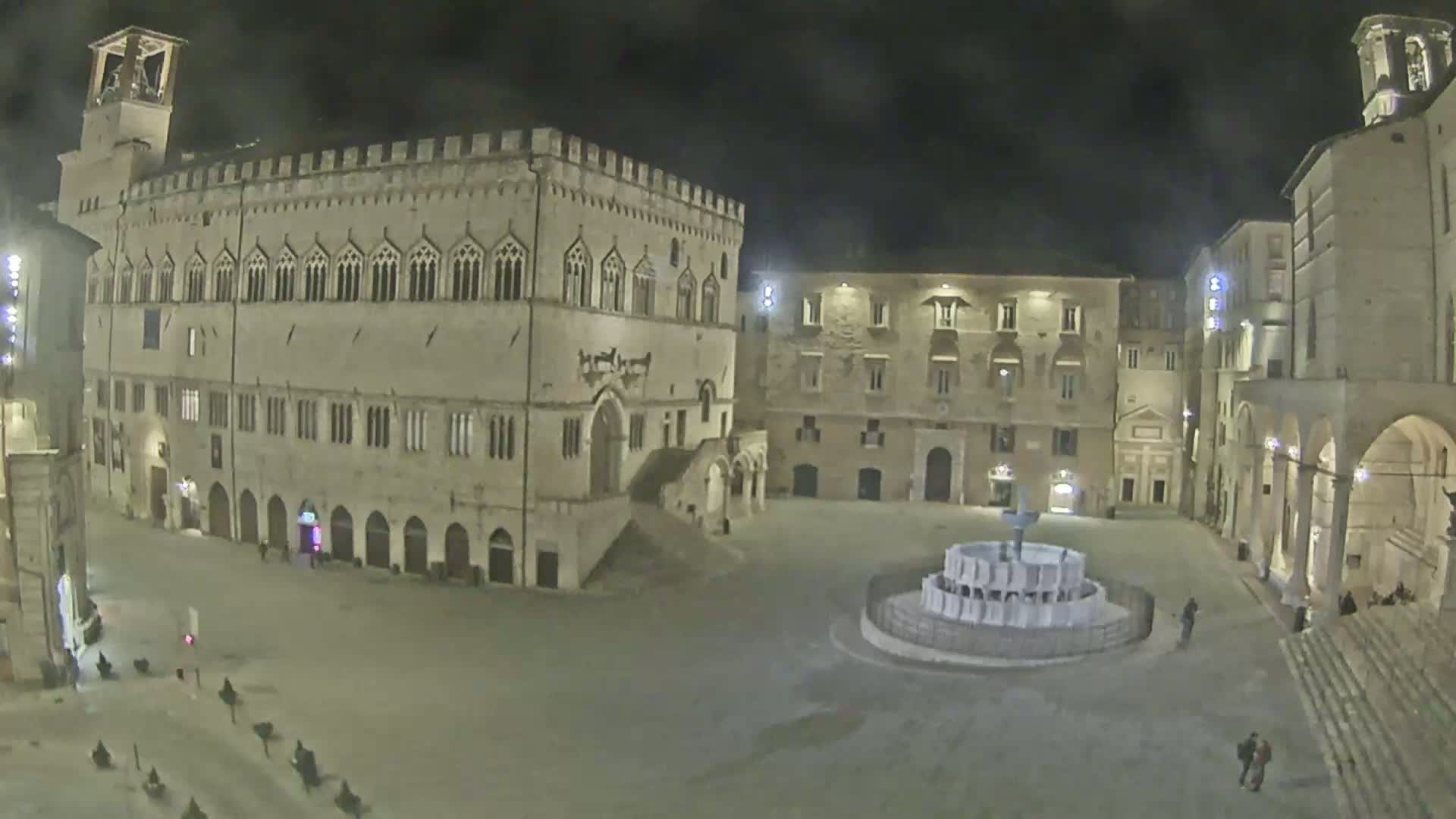 A mostly empty town square at night, featuring a large stone building, a fountain, and other historical structures under cloudy conditions.