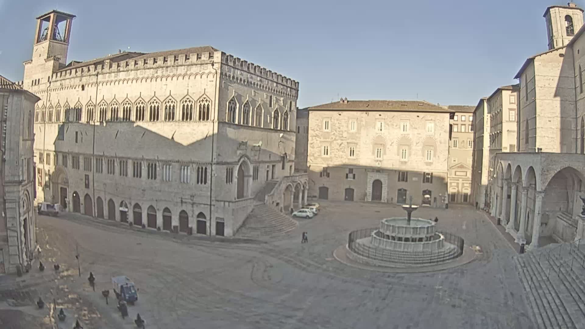 A large, mostly empty town square with a fountain at its center is surrounded by several stone buildings under a clear, sunny sky.