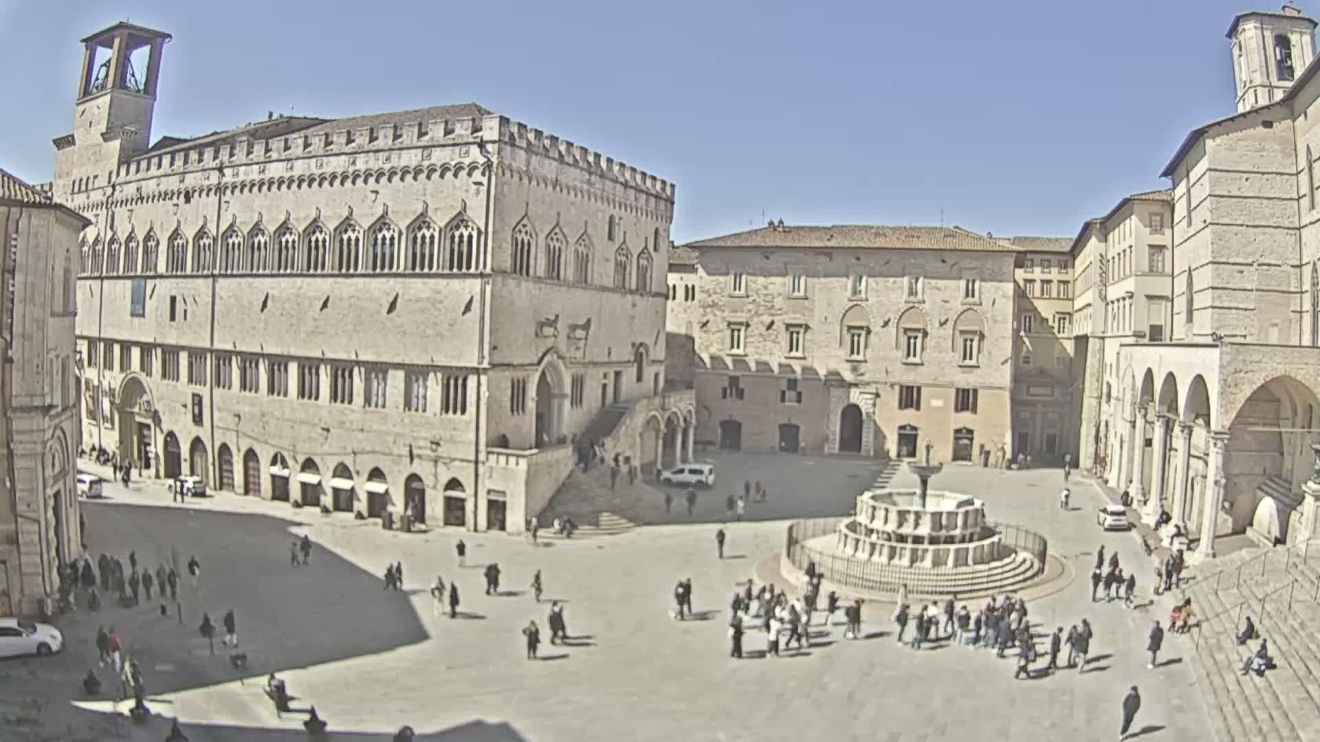 A large, sunny town square is surrounded by historic stone buildings, and features a fountain in the center.