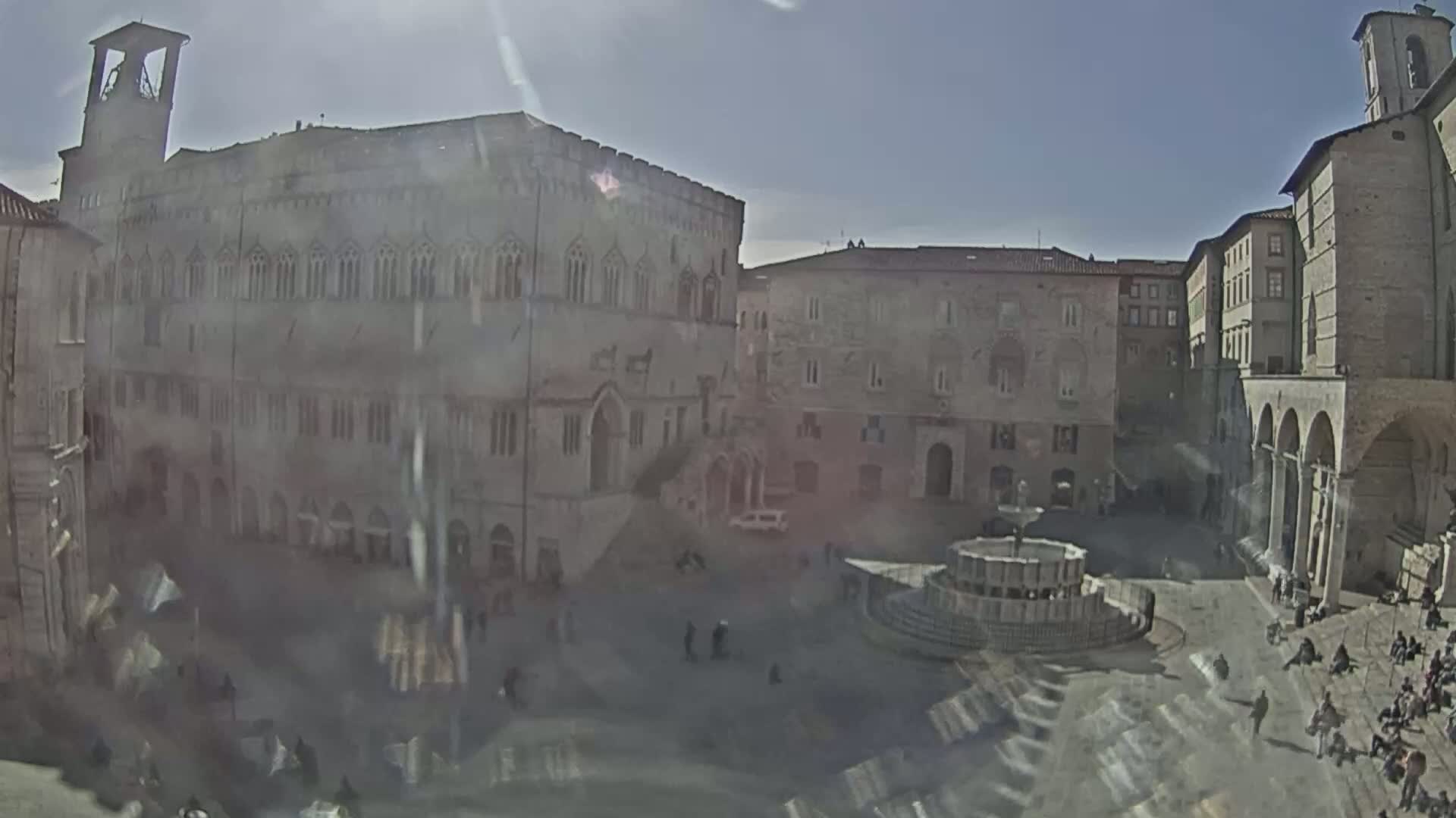 A sunlit town square with a central fountain, surrounded by stone buildings, some with arched walkways.