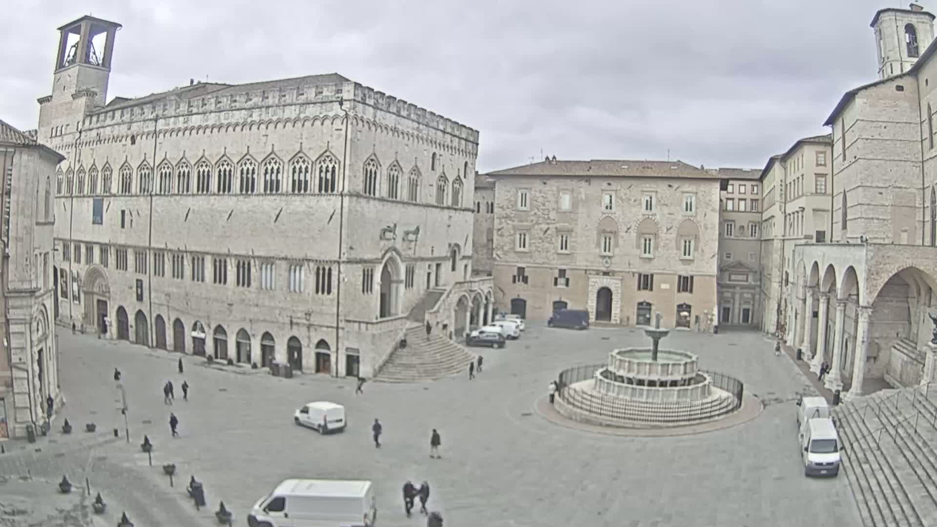 A large town square with several stone buildings, a fountain, and a few scattered people and vehicles under an overcast sky.