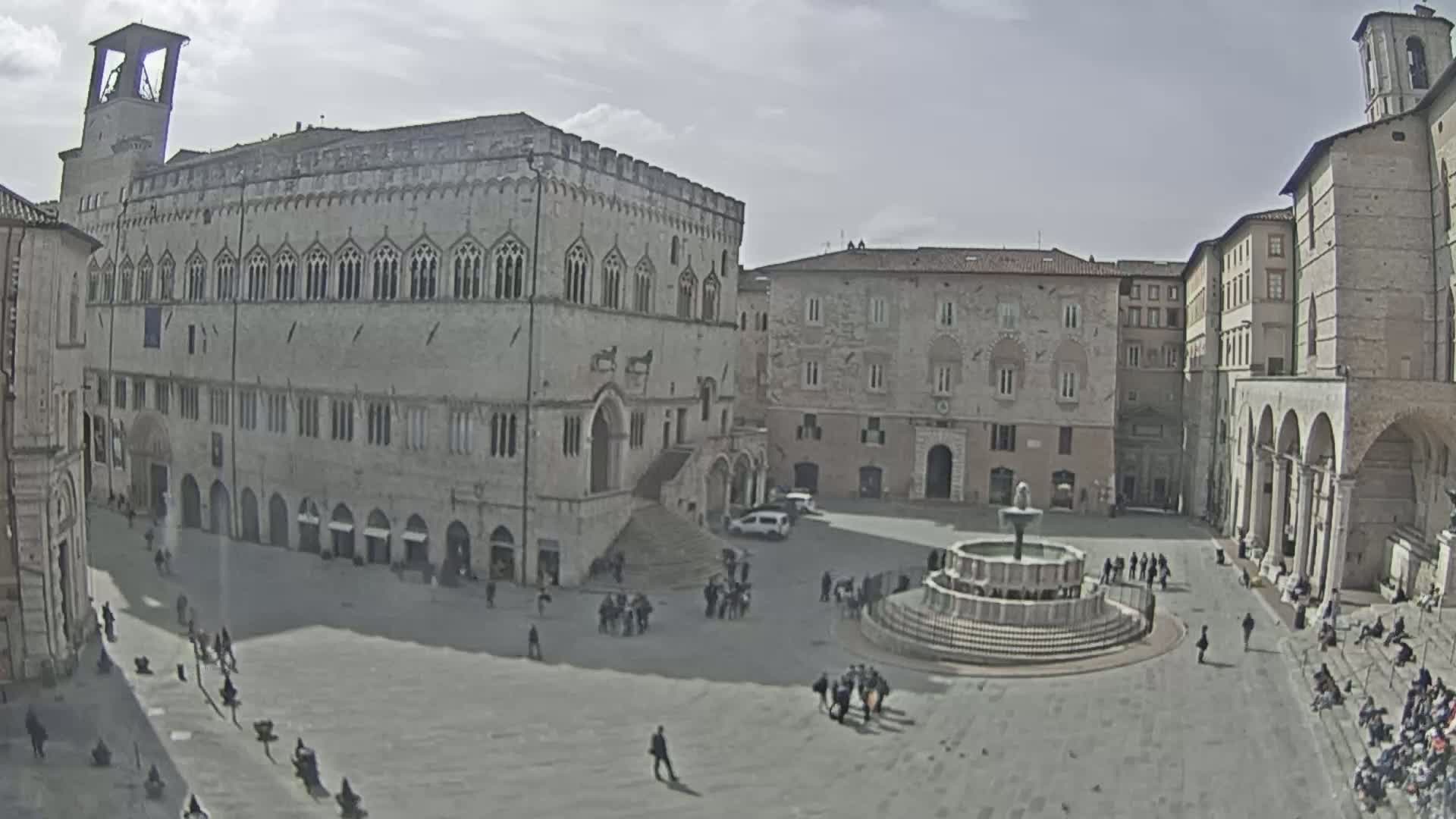 A large town square with a fountain in the center is surrounded by several historic stone buildings under a partly cloudy sky.