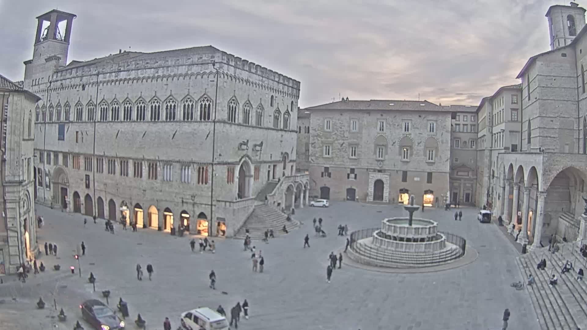 A large, stone plaza with a fountain at its center is surrounded by historic buildings under an overcast sky.