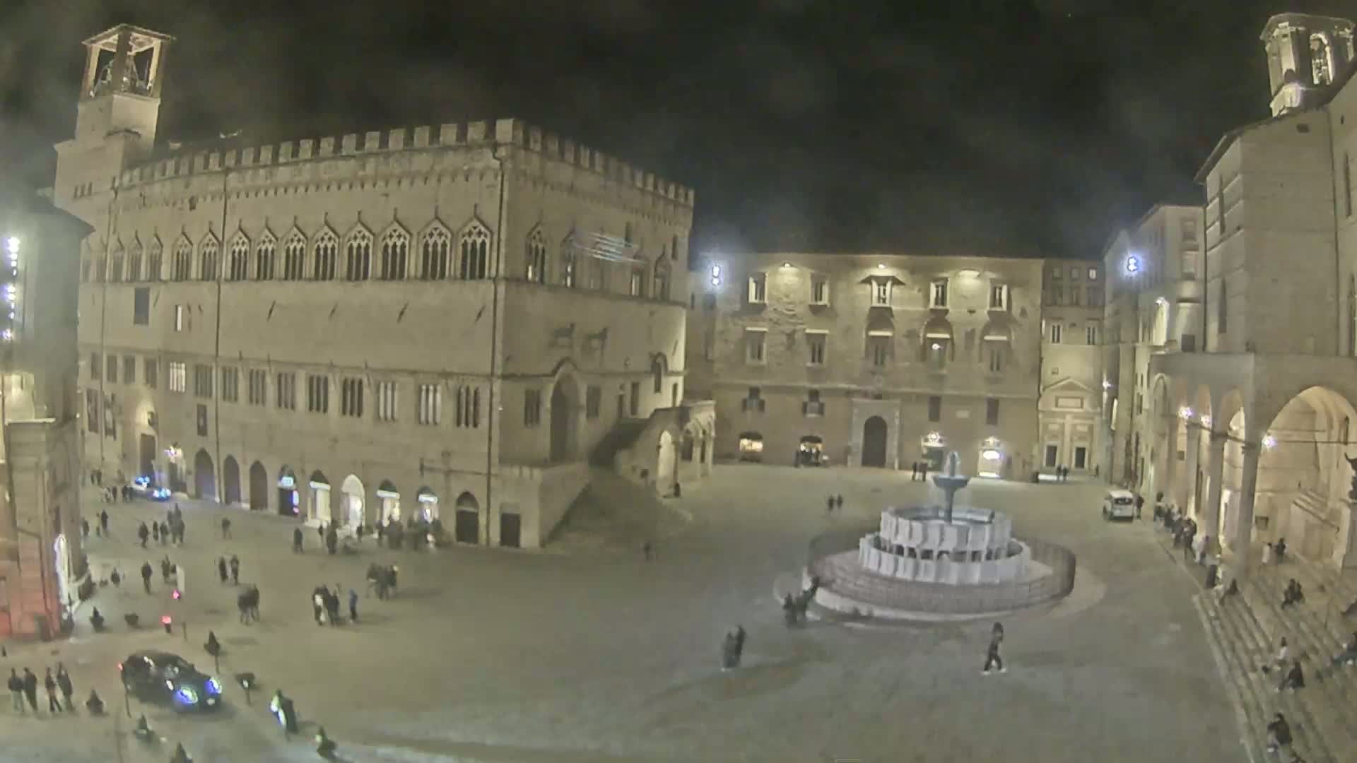 A nighttime view of a town square with a large stone building, several other buildings, a fountain, and a few people.
