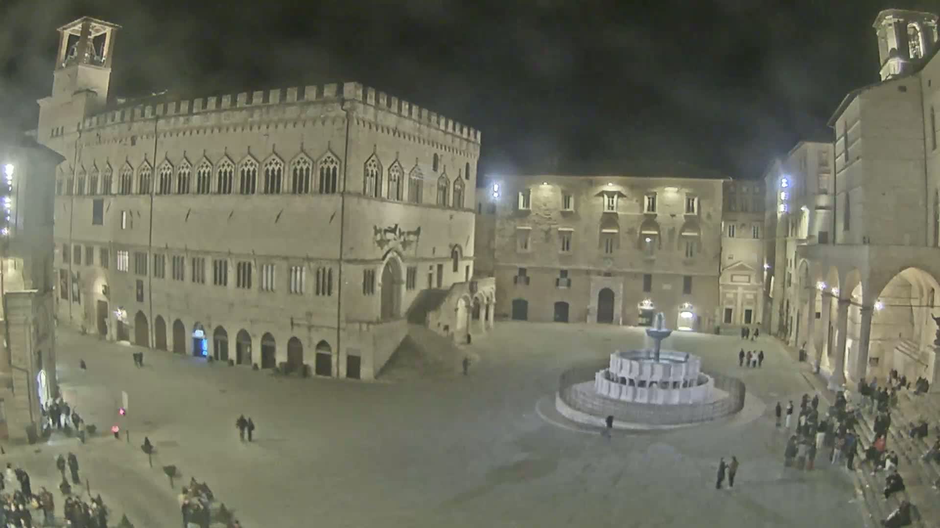 A nighttime view of a large town square with a fountain, surrounded by historic stone buildings under a cloudy sky.