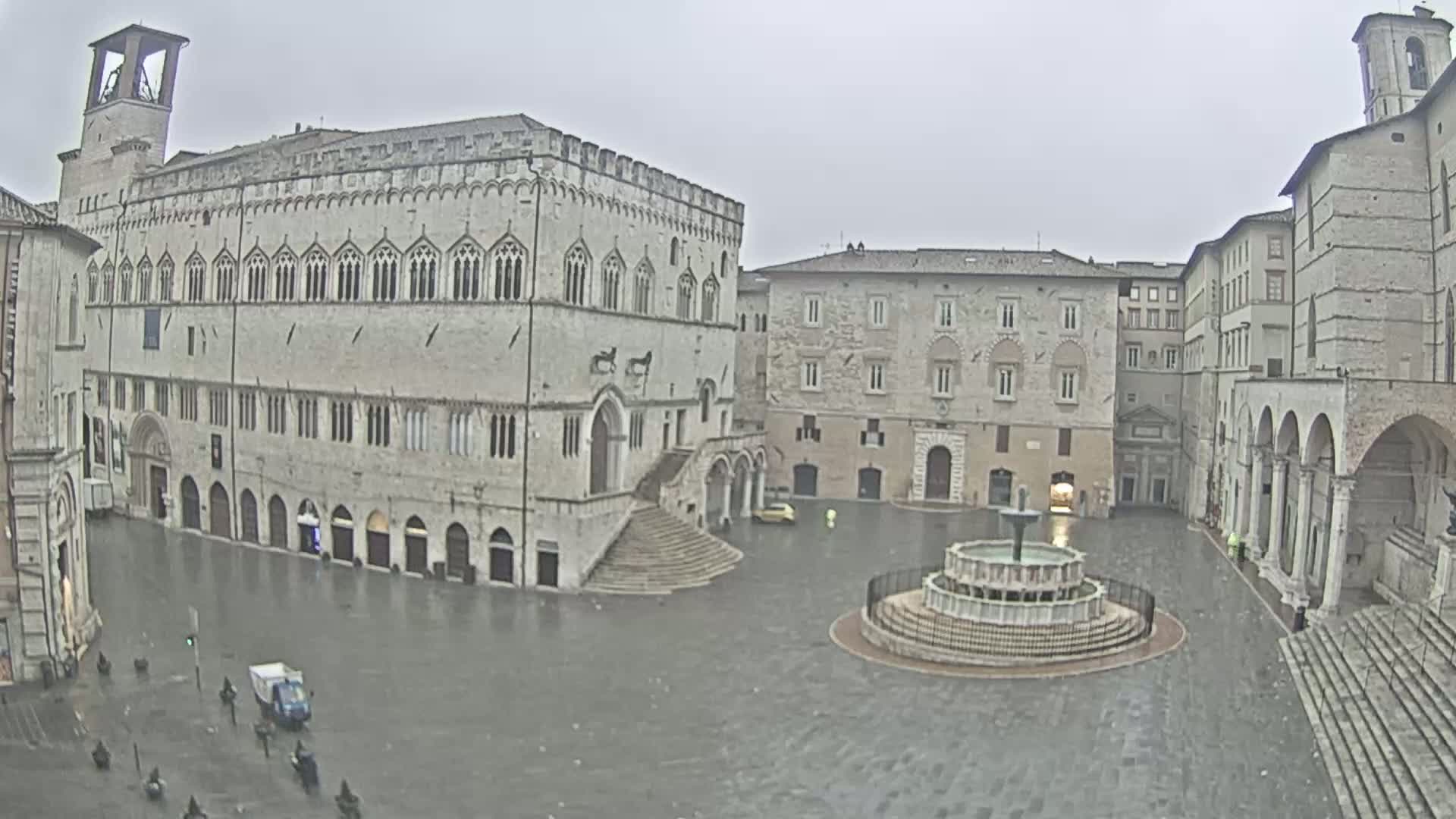 A large, stone plaza with a fountain in the center, surrounded by medieval-style buildings, on a rainy day.