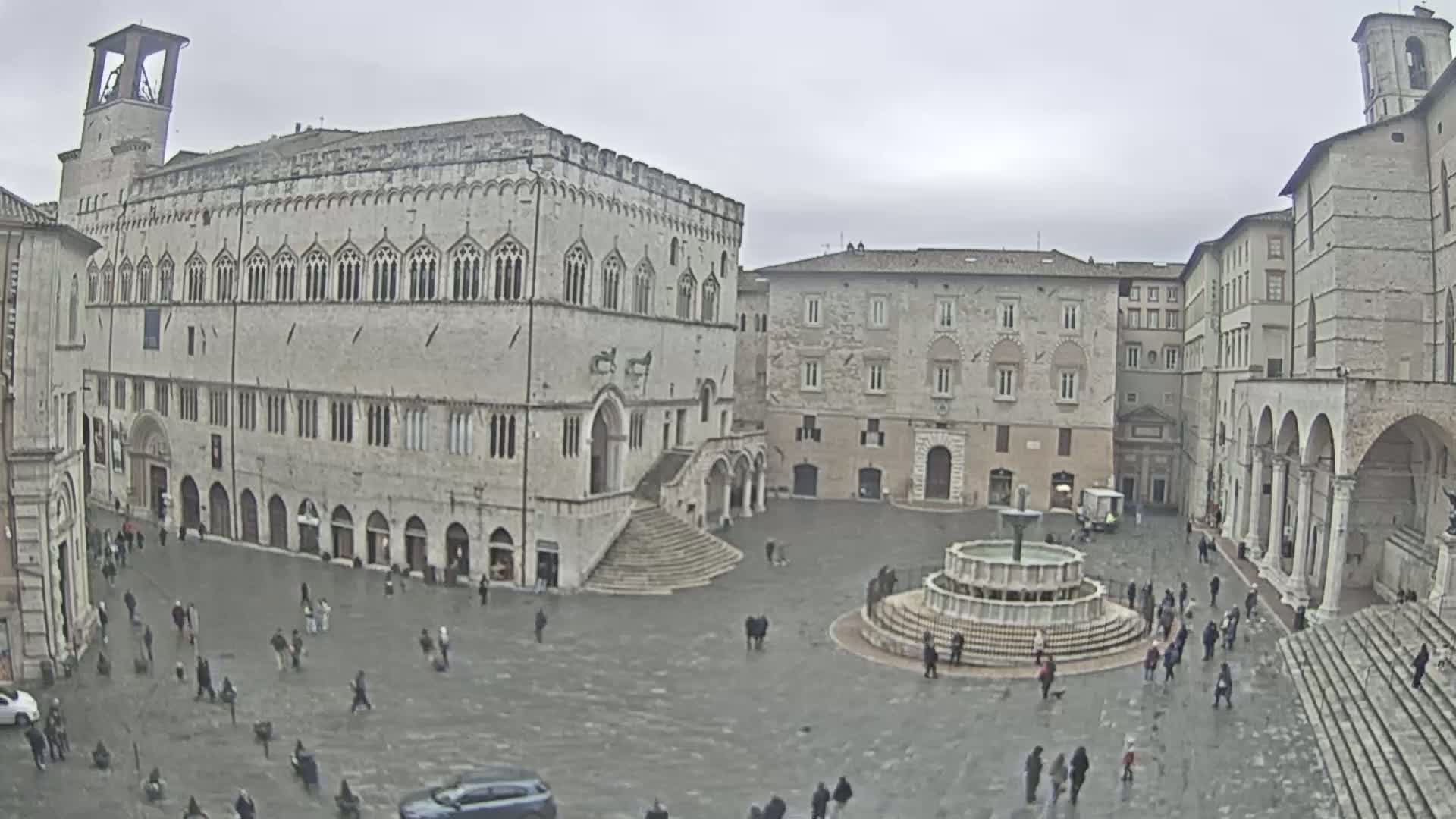 A large town square with several stone buildings, a fountain in the center, and a few people walking around under an overcast sky.