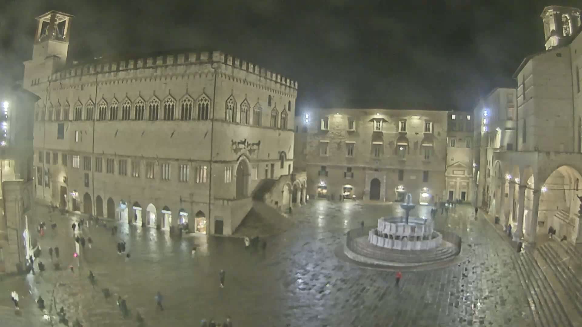 A rain-slicked town square at night, featuring a large stone building with arched entryways and a central fountain.