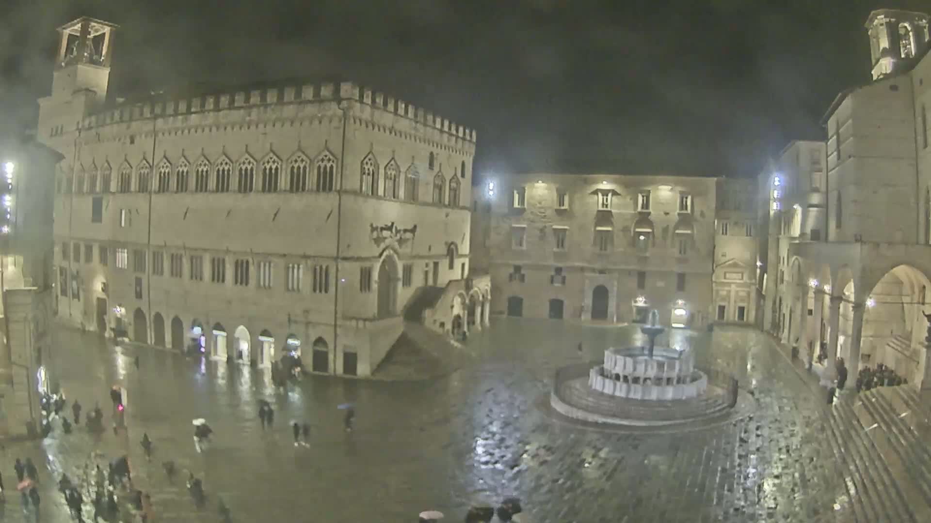 A rain-soaked town square at night, centered around a fountain, is surrounded by historic stone buildings.