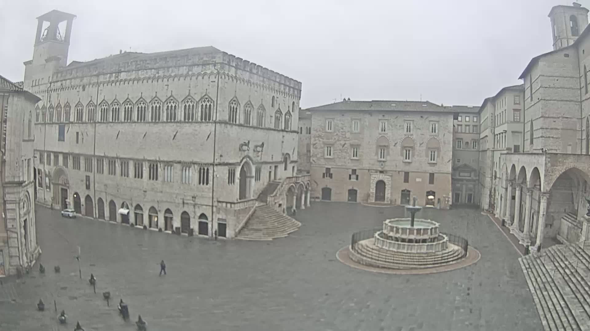A large, empty stone plaza with a fountain in the center, surrounded by medieval-style buildings on a foggy day.