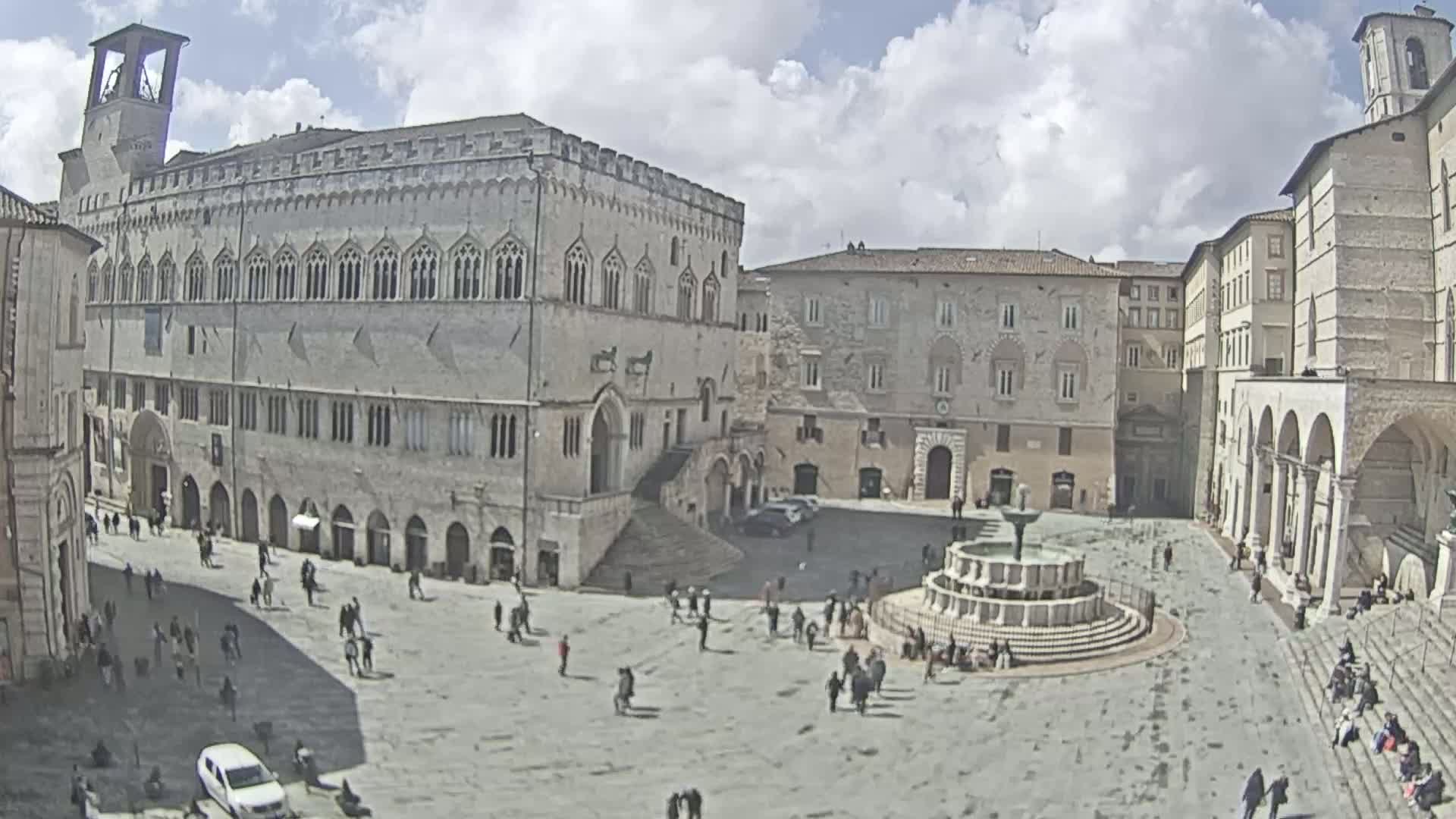 A large town square with a fountain and several stone buildings is seen on a partly cloudy day.