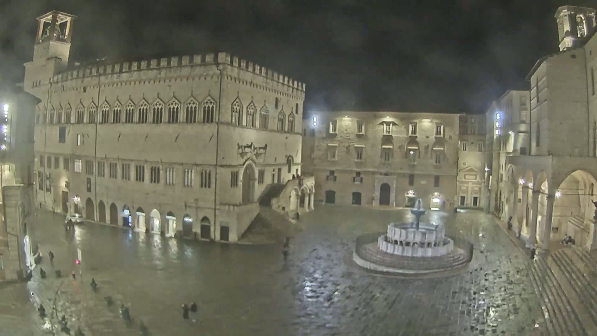 A large, stone plaza at night, with a fountain in the center and several buildings surrounding it, is flooded with rainwater.