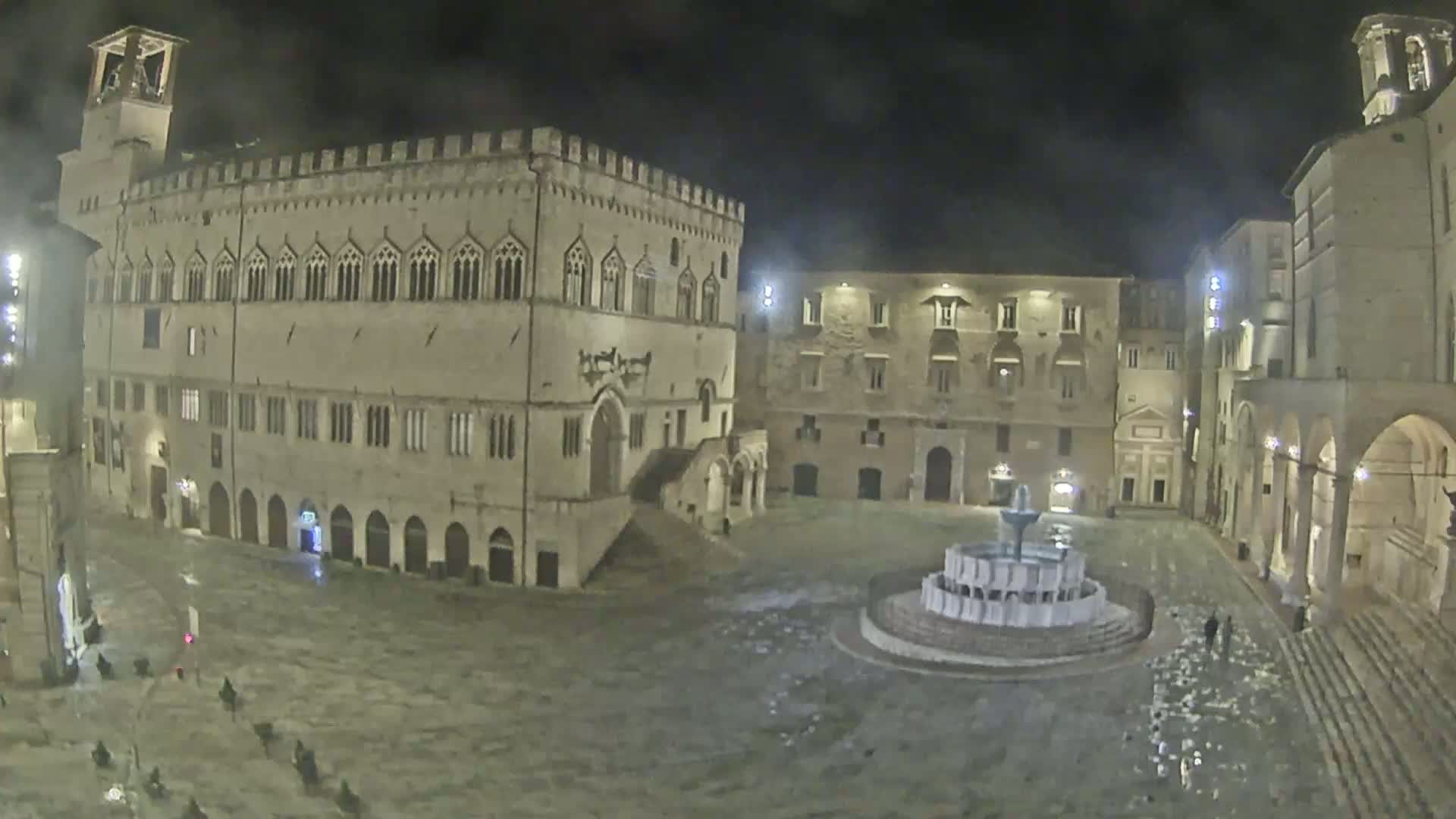 A nighttime view of a large, stone plaza with a fountain at its center, surrounded by medieval-style buildings.