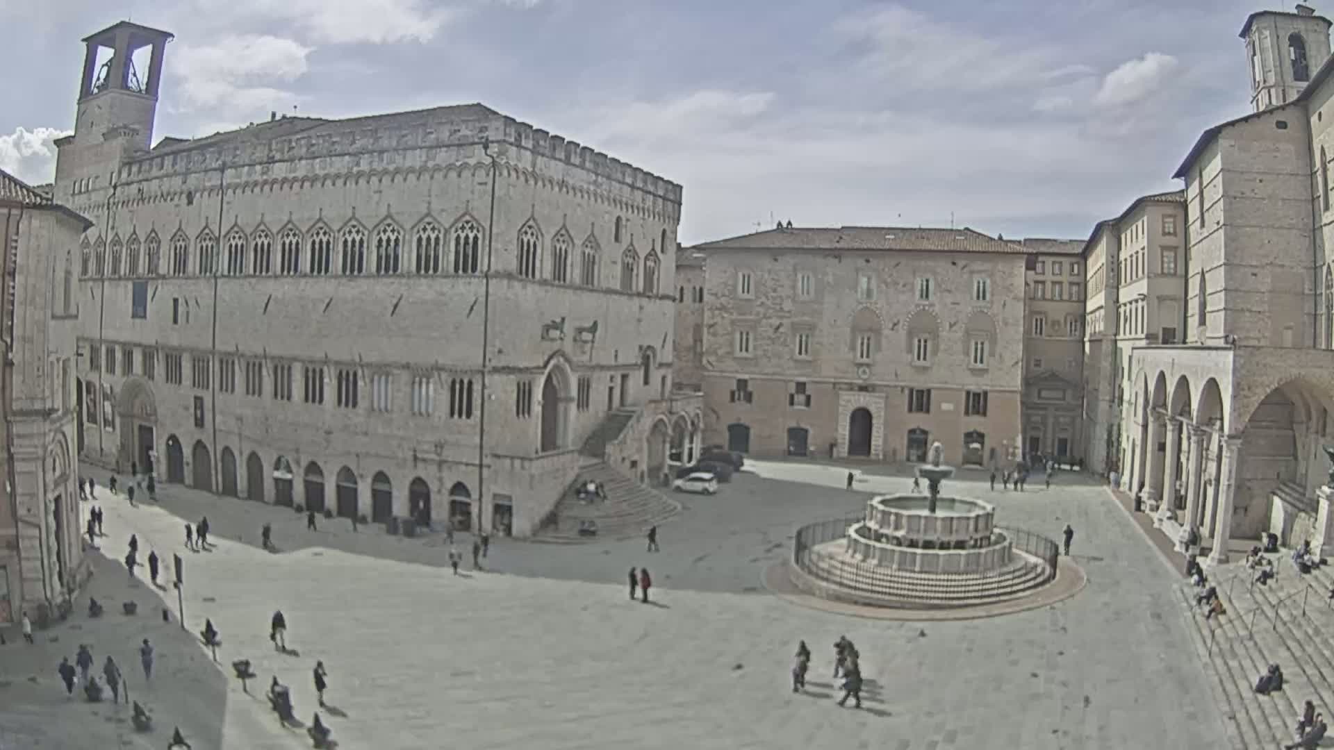 A large, mostly empty town square with a fountain, surrounded by several stone buildings under a partly cloudy sky.
