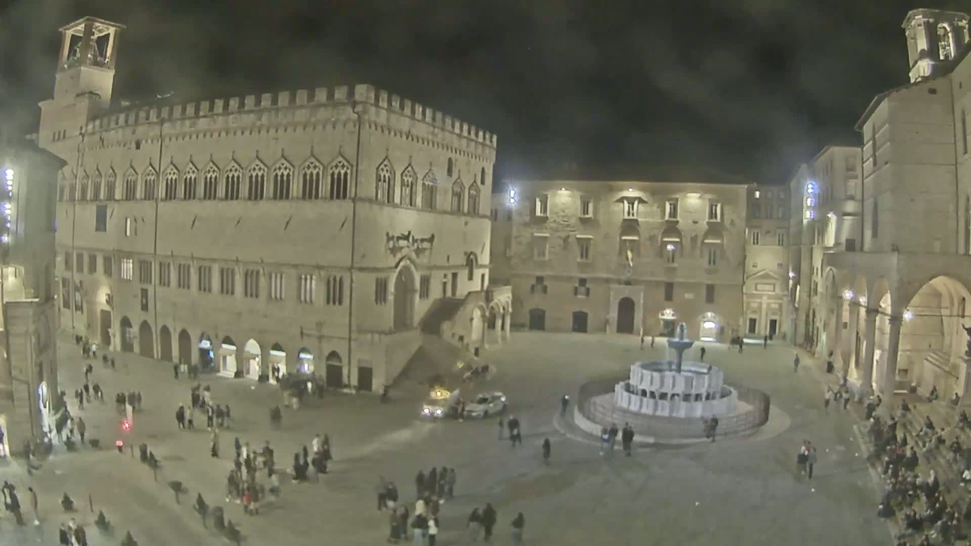 A nighttime view of a large town square with a fountain at its center, surrounded by historic stone buildings and many people.