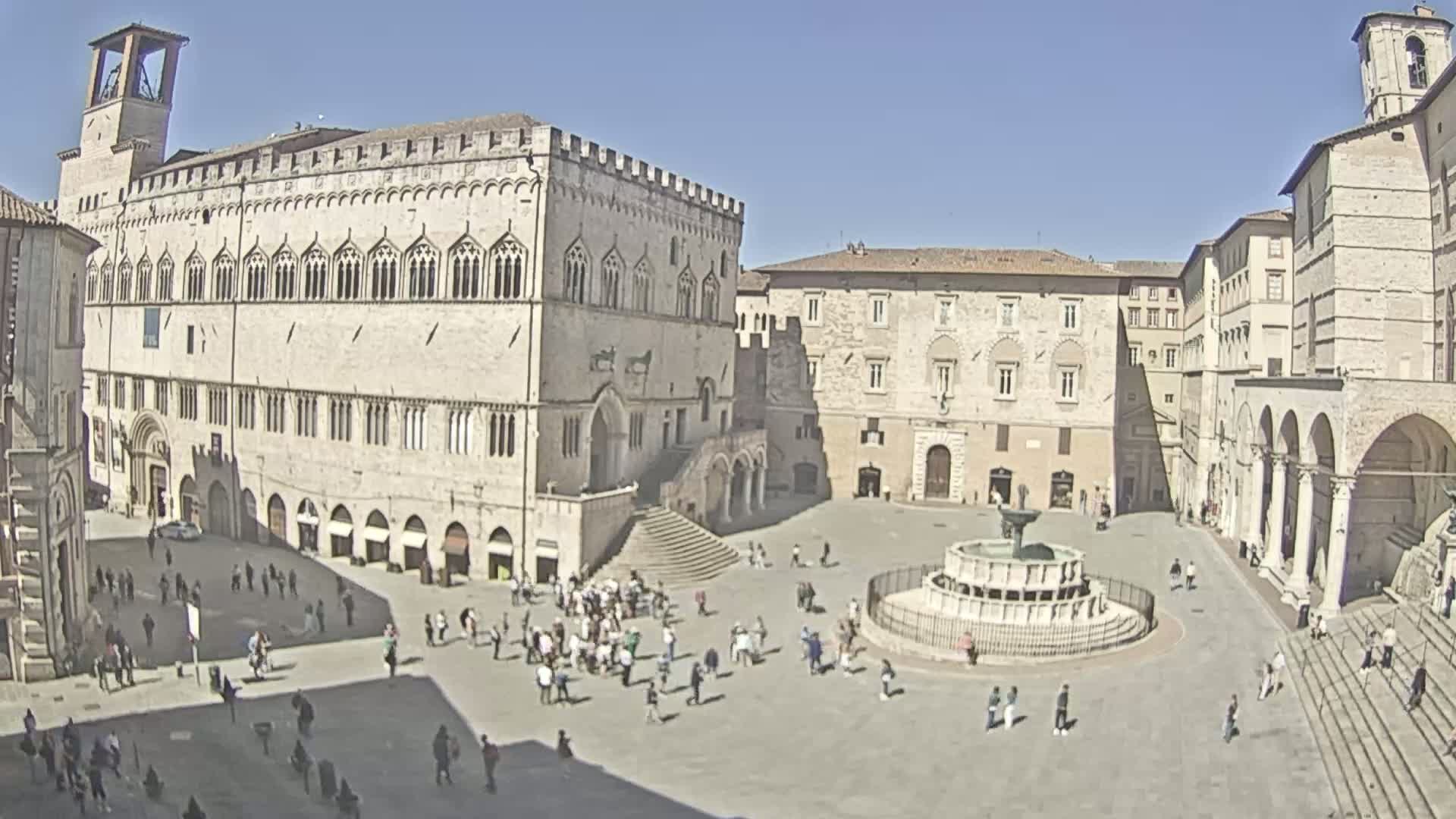 A large, light-colored stone plaza, under a sunny sky, is surrounded by historic buildings including a grand palace with a staircase, and features a fountain at its center, with many people scattered throughout.