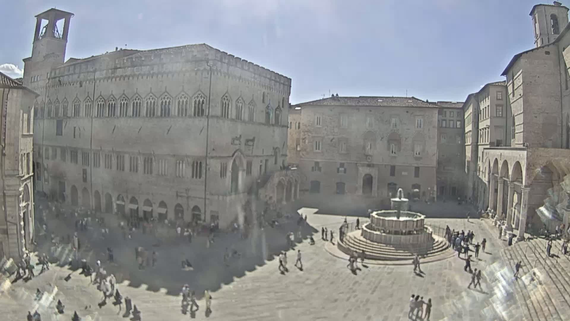 A sunny town square is filled with people, centered around a tiered fountain, with large stone buildings surrounding it.