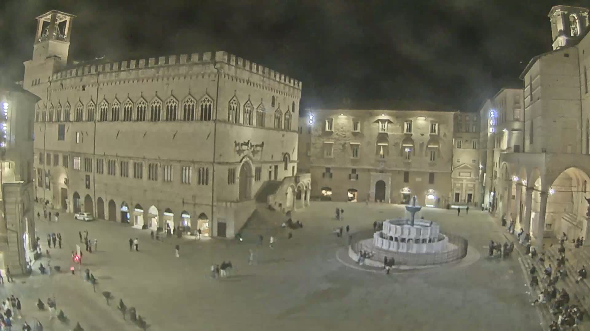 A nighttime view of a town square with historical buildings, a fountain, and a few people scattered throughout.