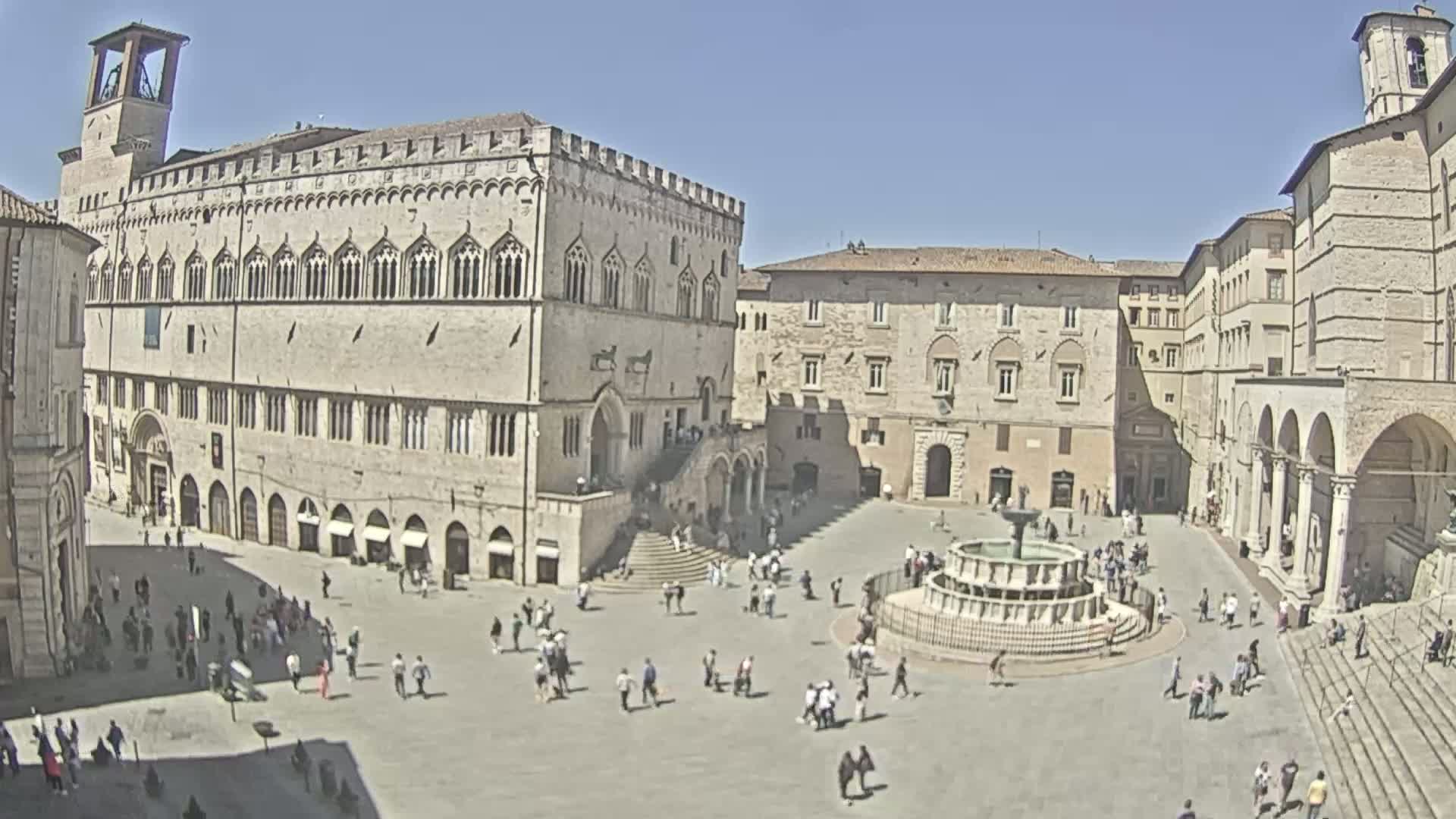 A large, sunny town square is filled with people and features a central fountain, surrounded by historic stone buildings.