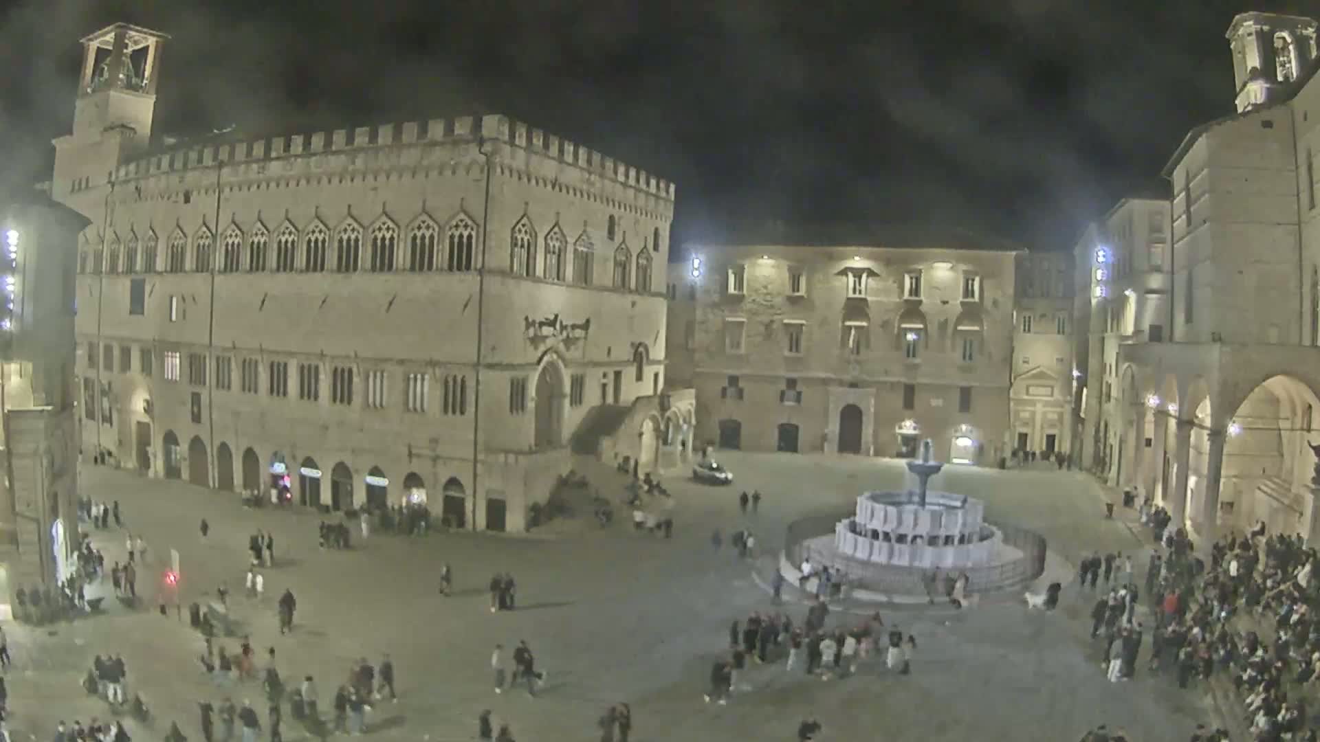 A nighttime, outdoor scene shows a large town square with a fountain in the center, surrounded by several historic buildings and many people.